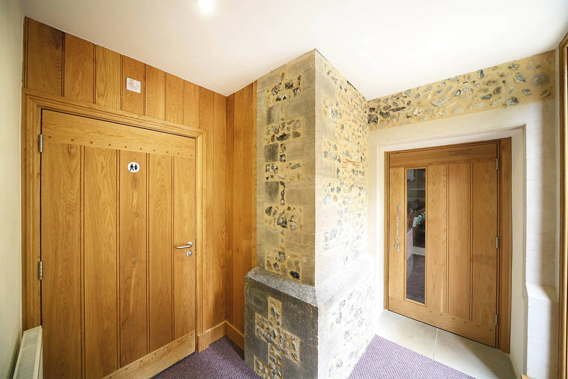 Interior hallway with wooden doors, stone and brick wall, and a small rectangular window in the door on the right side.