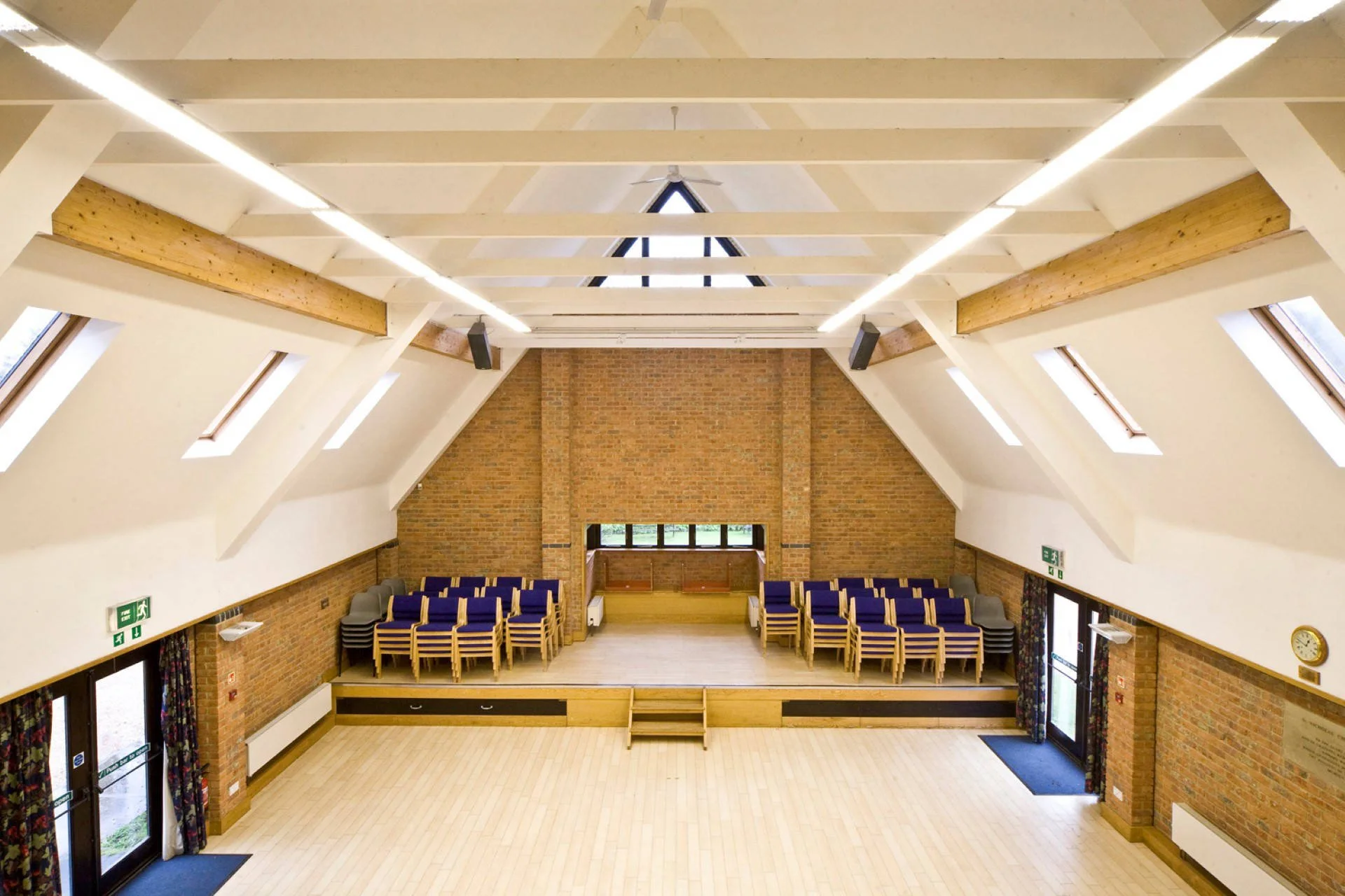Large conference room or hall with wooden floors, a high ceiling with exposed wooden beams, skylights, and wall-mounted speakers.