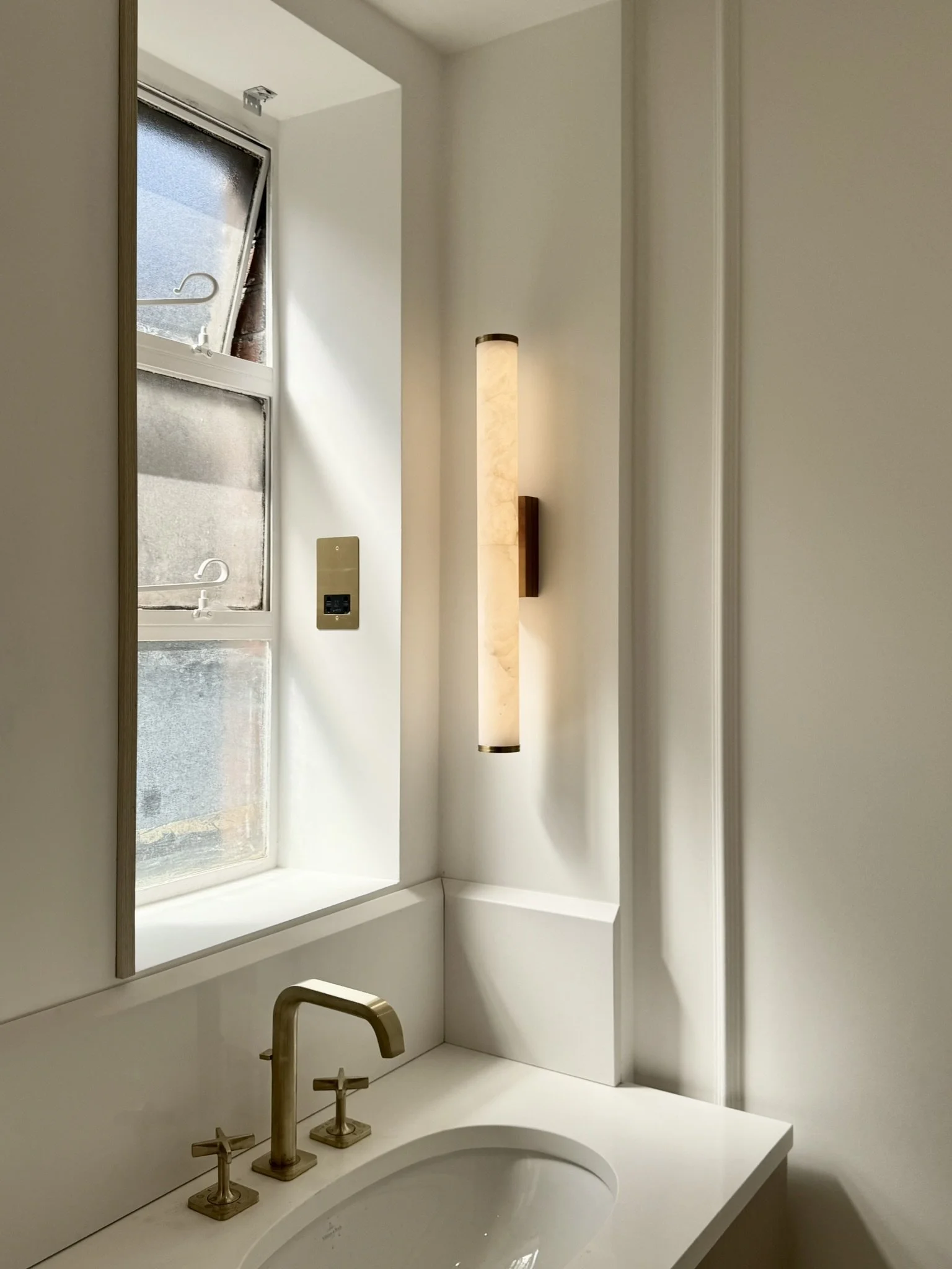 A modern bathroom sink with gold-toned fixtures, a rectangular mirror, a beige wall-mounted light fixture, a window with frosted glass, and a beige wall with crown molding.