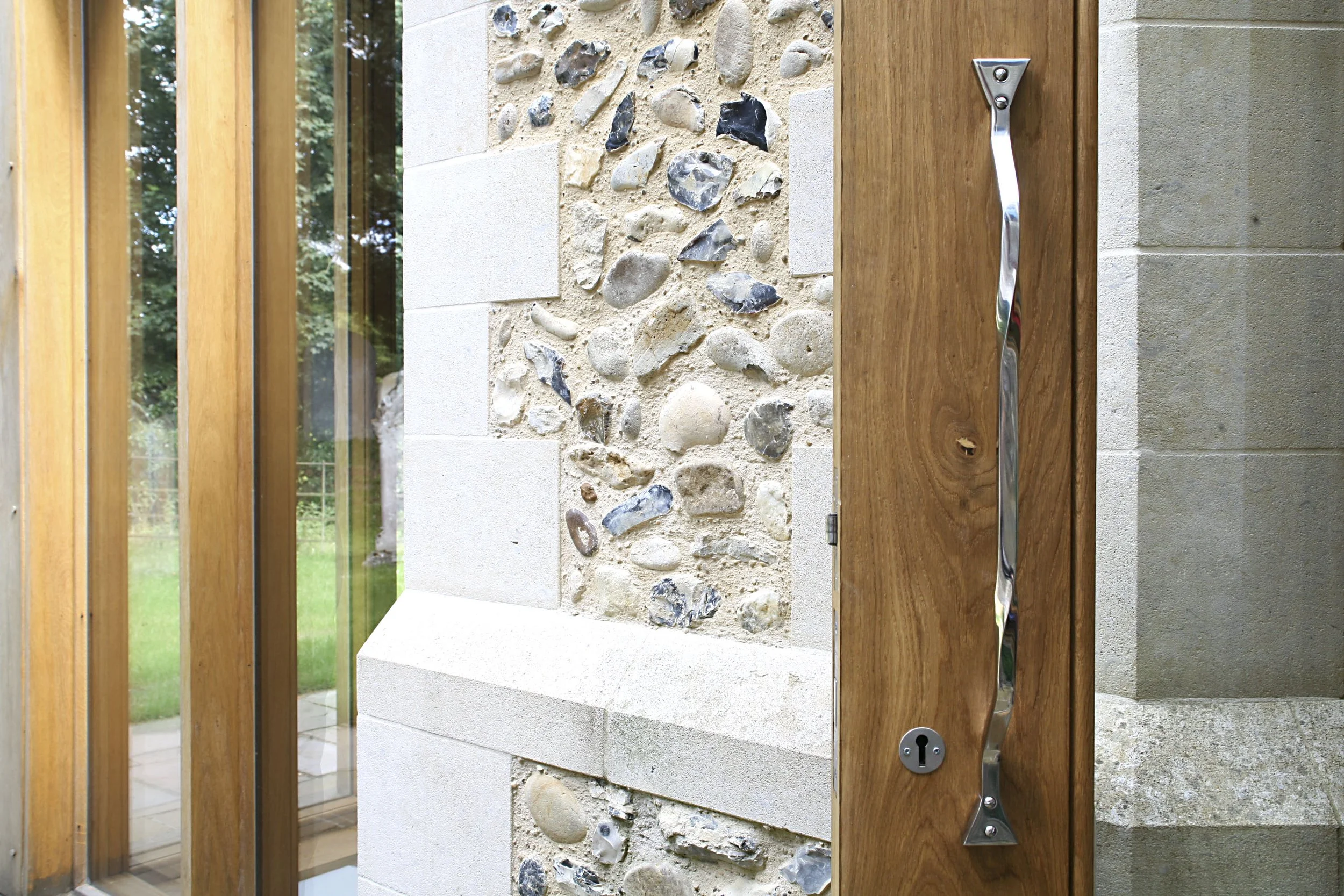 Close-up of a modern wooden front door with a metal handle and a security lock, next to a wall with decorative pebble and plaster finish, and a window with glass panels reflecting surrounding greenery.
