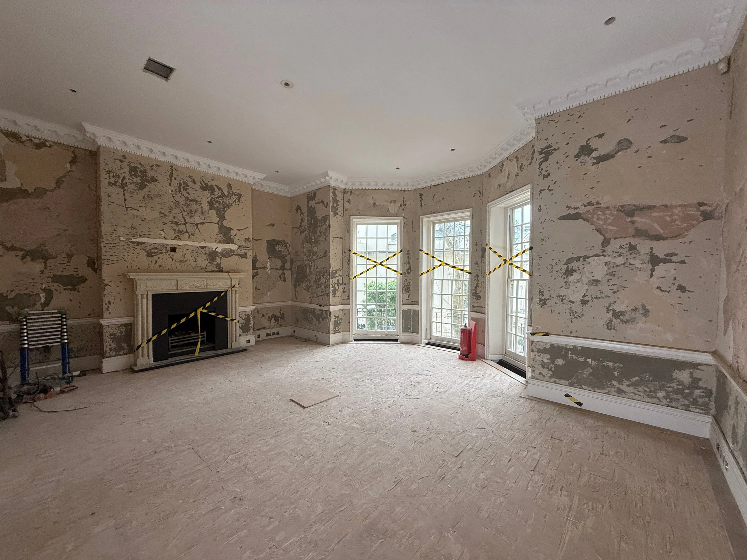A spacious living room under renovation with walls stripped of paint or wallpaper, revealing plaster and drywall. The room has high ceilings with intricate crown molding, large windows letting in natural light, and a fireplace with decorative molding