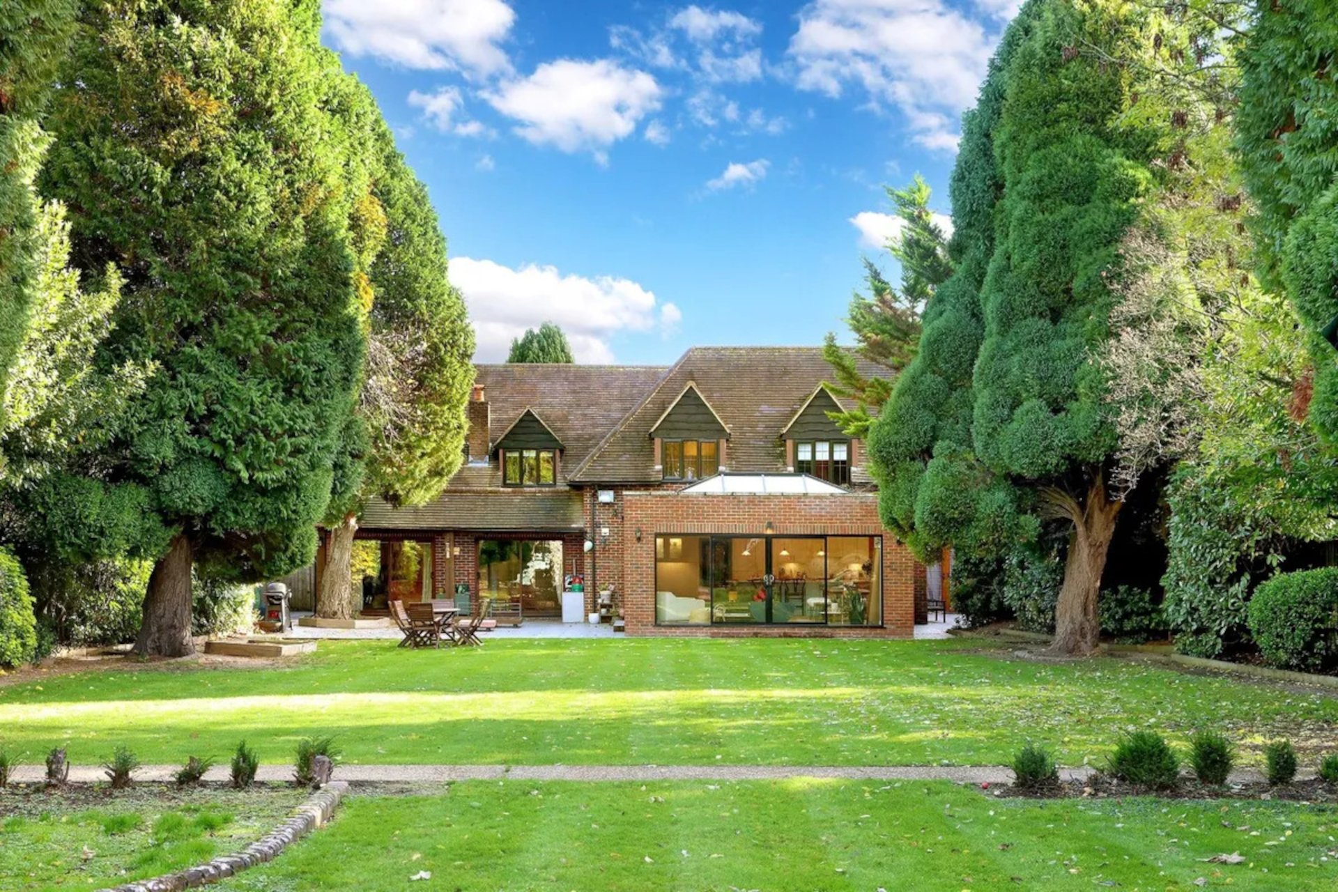 A large brick house with a sloped roof, surrounded by tall, lush green trees and an outdoor patio area with a table and chairs, on a sunny day with a blue sky and white clouds.