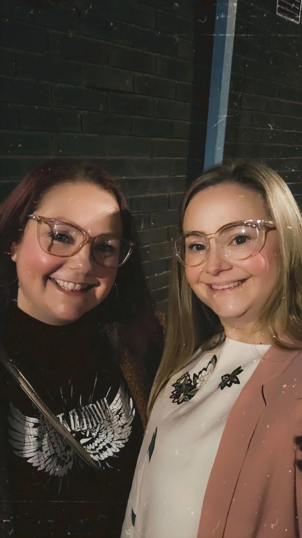 Two women with glasses smiling and posing for a photo against a dark brick wall.