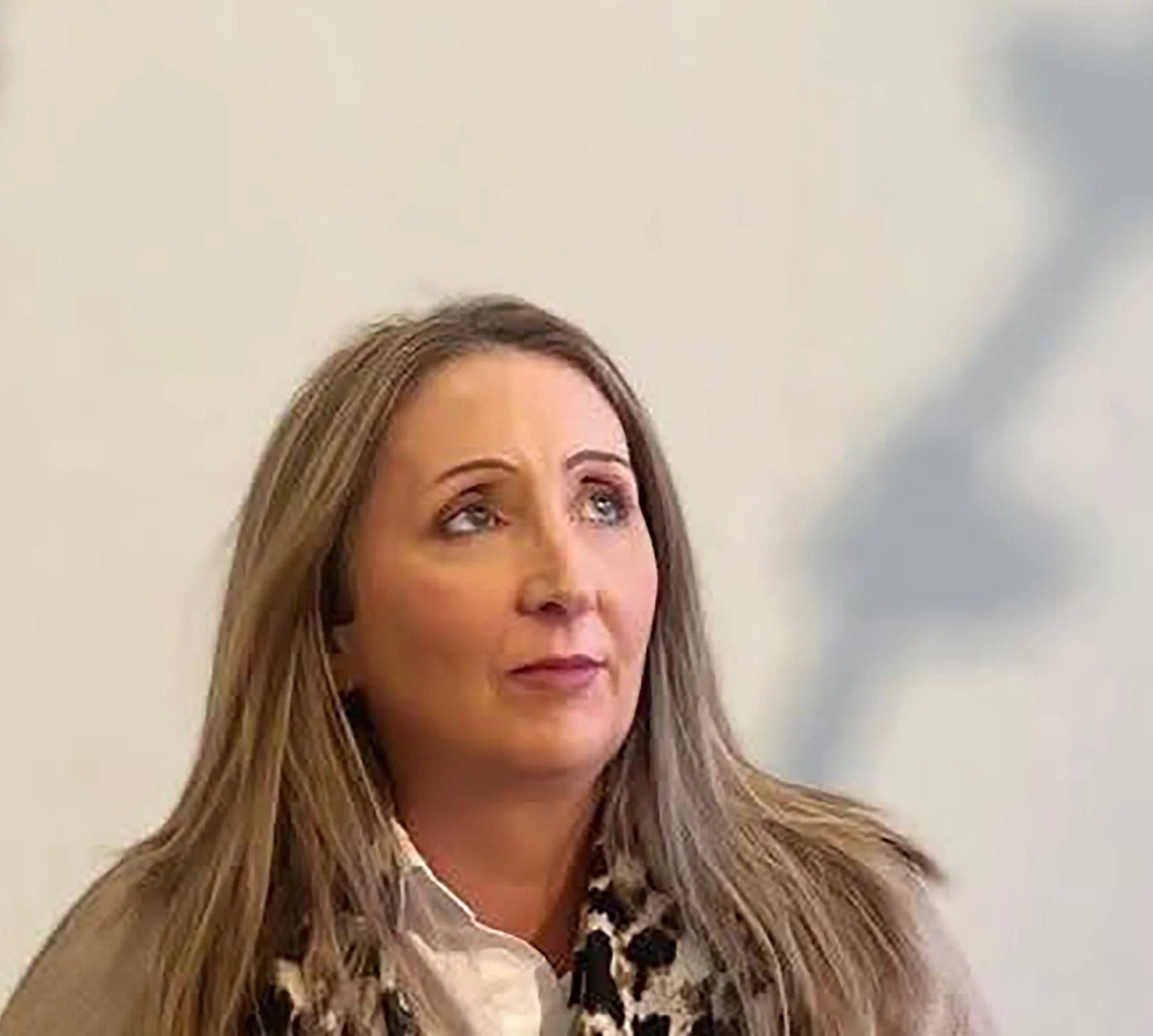 A profile picture of Suzanne Buckley , wearing a beige sweater and a black and white patterned scarf, looks upwards with a thoughtful expression against a plain light-colored wall.