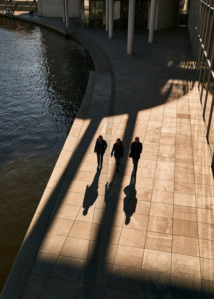 Three people walking along a waterfront promenade, with their shadows cast on the ground by the sunlight, alongside modern buildings.