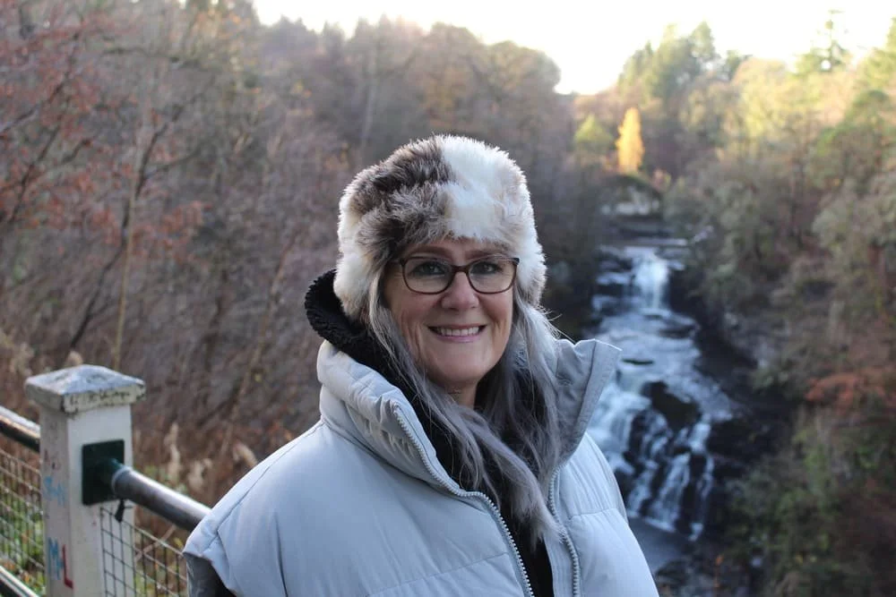 Woman in a warm jacket and furry hat smiling outdoors near a waterfall in a forested area.