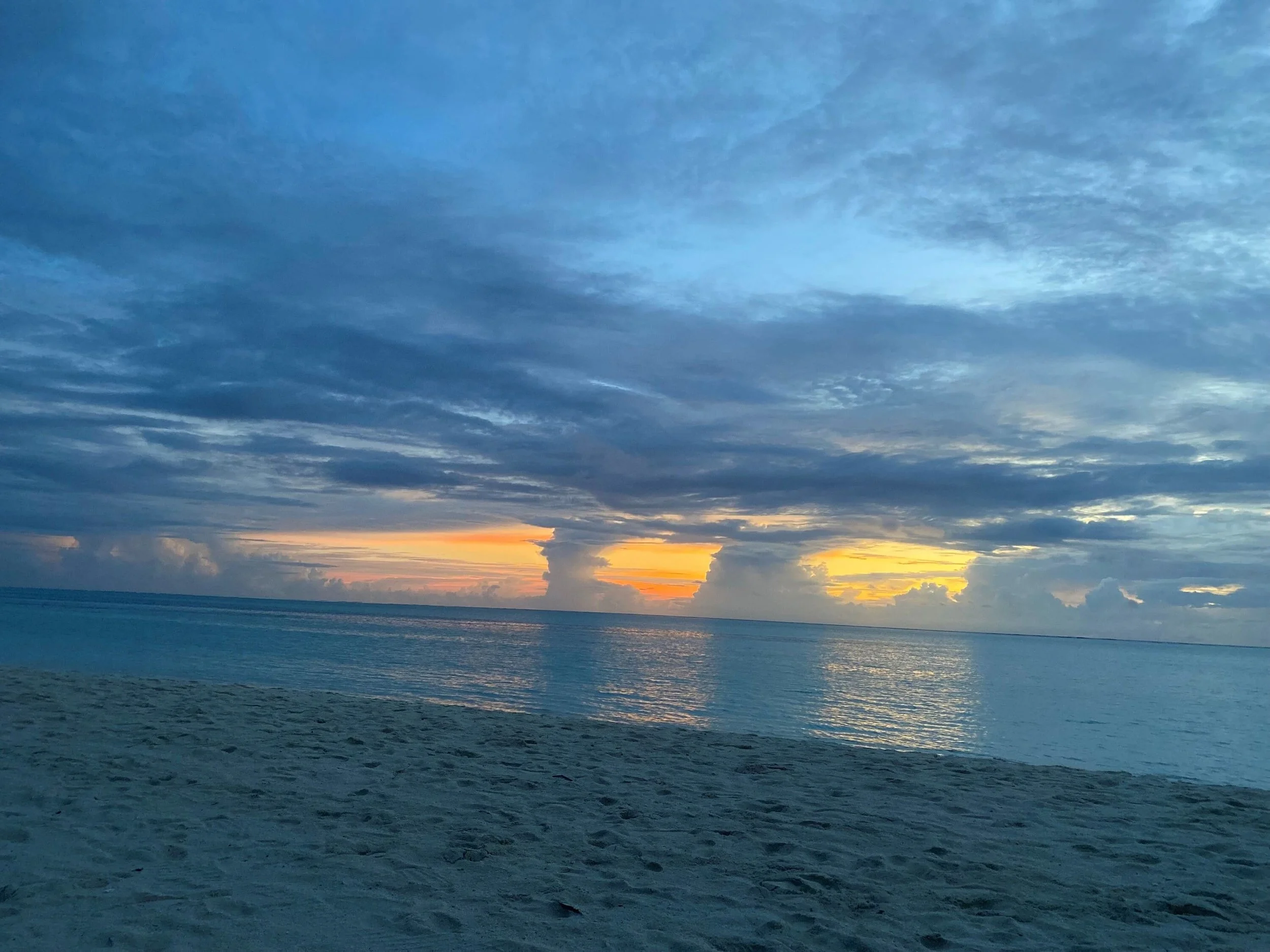 Sunset over the ocean with blue sky and scattered clouds, sandy beach in the foreground.