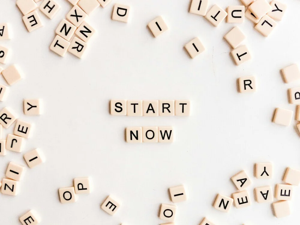 Scrabble tiles spelling out "START NOW" surrounded by scattered tiles with letters on a white background.