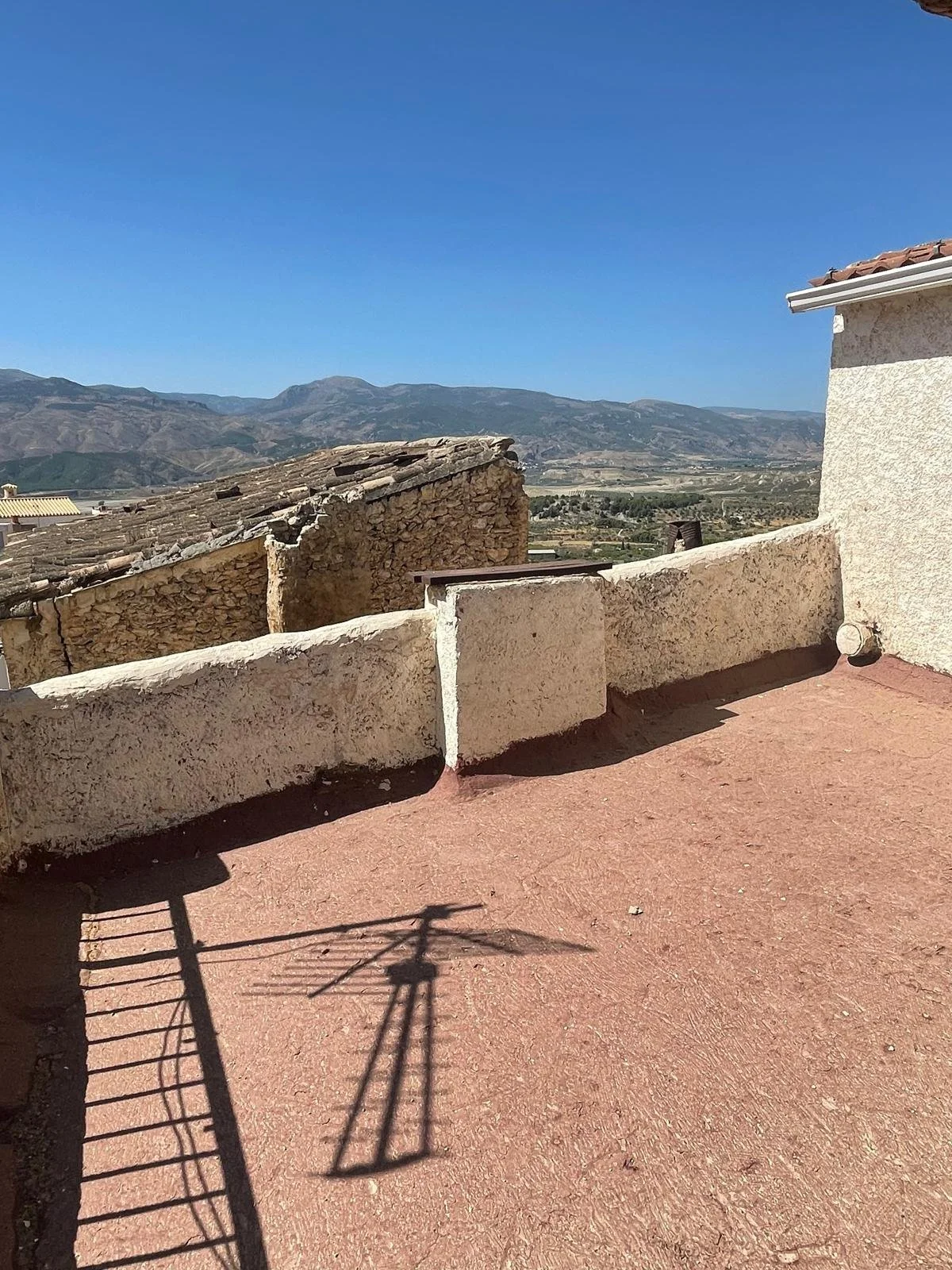 View of a rooftop terrace with a red concrete floor, beige stucco walls, and a metal railing casting a shadow. In the background are mountains and a clear blue sky.