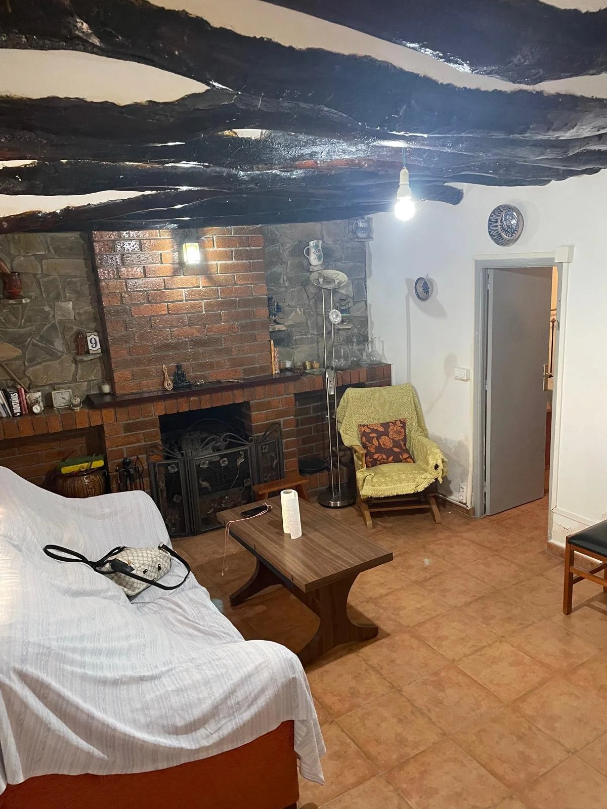 Living room with a brick and stone fireplace, white sofa with a small bag on it, dark brown coffee table with a roll of paper towels, yellow armchair with a floral pillow, standing lamp, and doorway in the background.