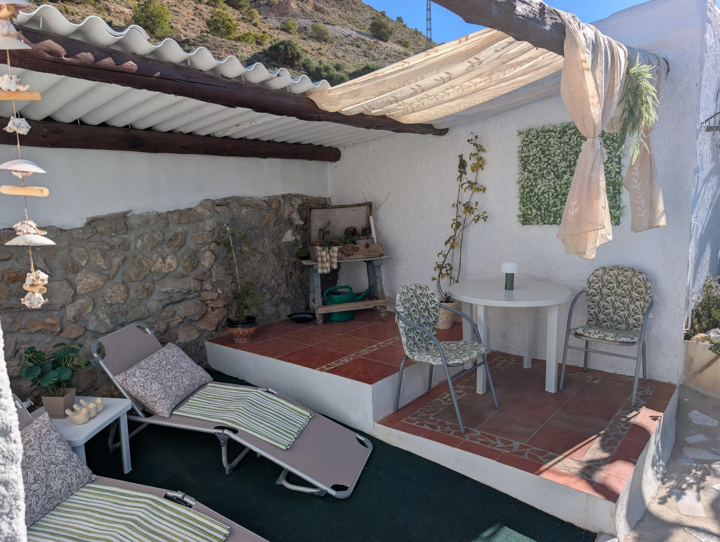 Outdoor patio with white stucco walls, terracotta tile flooring, and a stone accent wall. Features a small round table with two chairs, a lounging chair with pillows, a shaded area with a draped fabric canopy, and potted plants. There's a hillside with sparse bushes in the background.