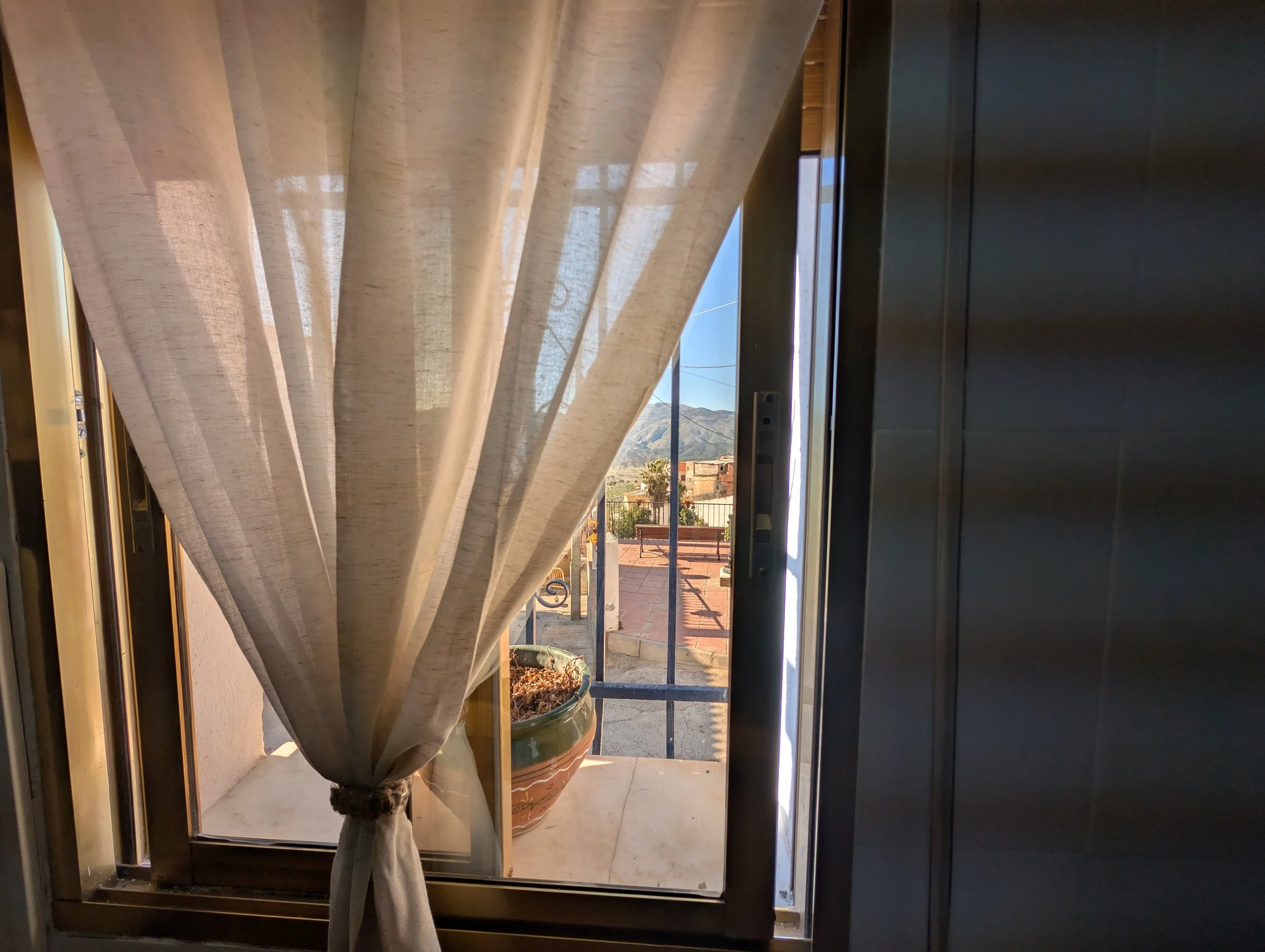 View through an open window with beige curtains, showing a small balcony with a large potted plant, a bench, and mountains in the background.