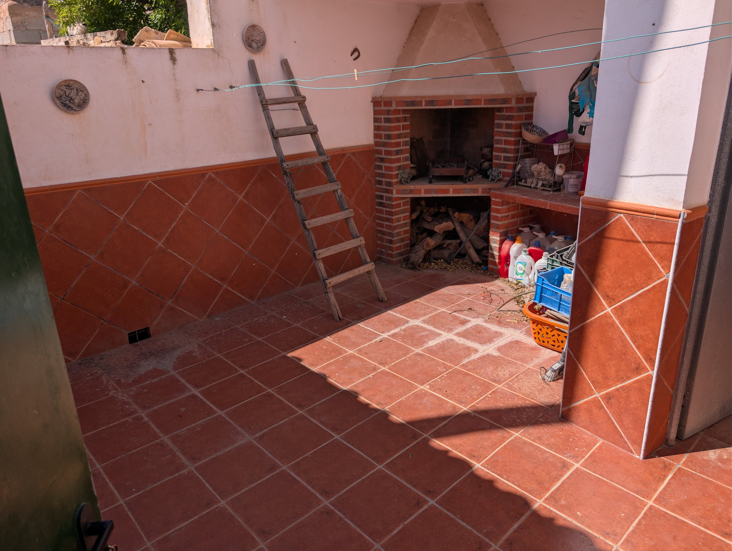 Outdoor patio with brick fireplace, logs stored below, with cleaning supplies and containers on the right, a wooden ladder leaning against the wall, and a clothesline overhead, partially shaded.
