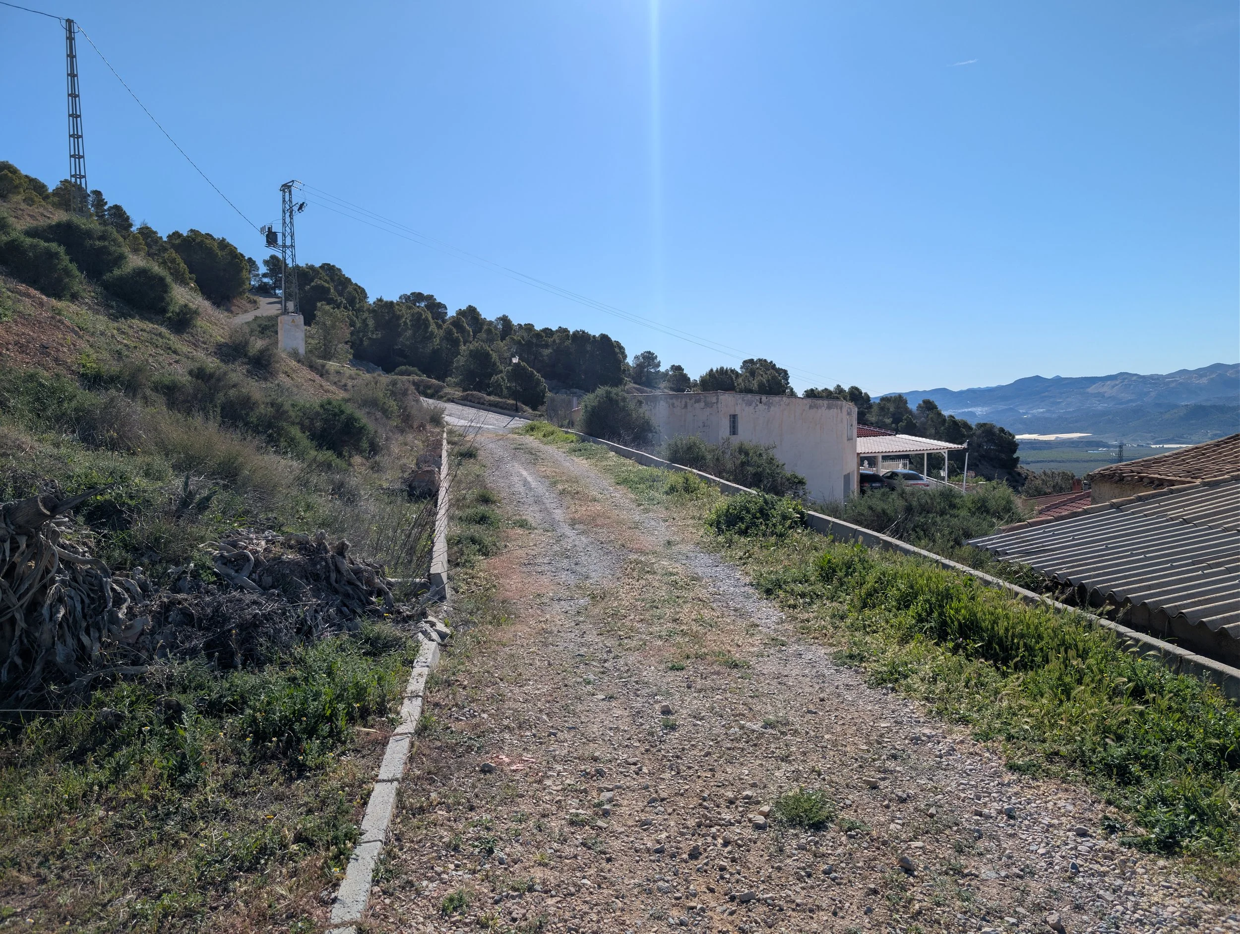 A dirt road along a hillside with houses and mountains in the background, under a clear blue sky.