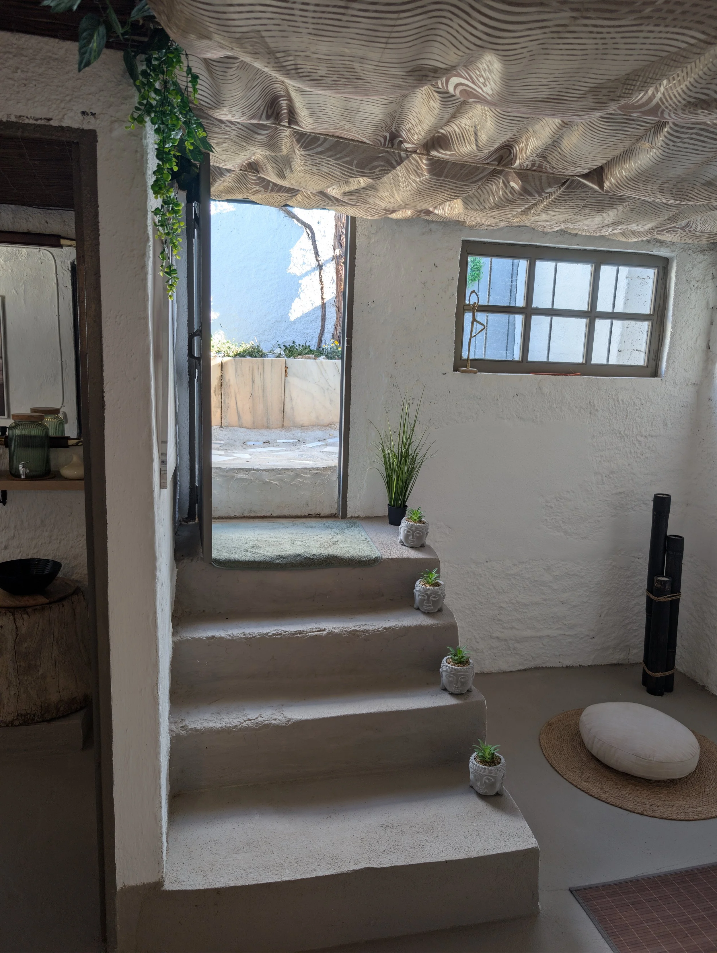 Interior of a cozy room with white textured walls, concrete stairs leading outside, and a small window. Decor includes potted plants, a round cushion on a woven mat, and fabric ceiling covering.