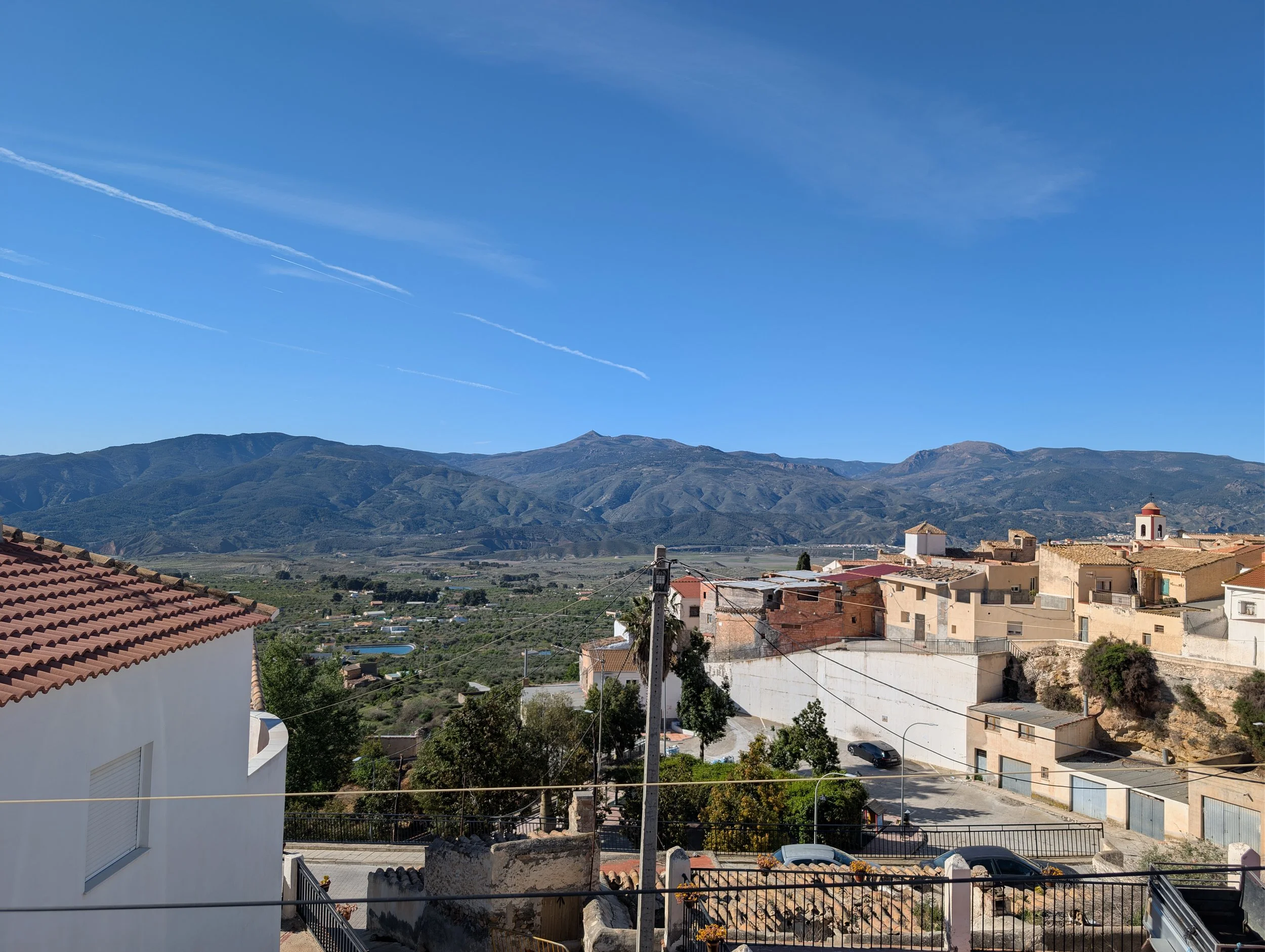 View of a hillside town with houses, trees, and streets, set against mountain ranges under a clear blue sky with contrails.