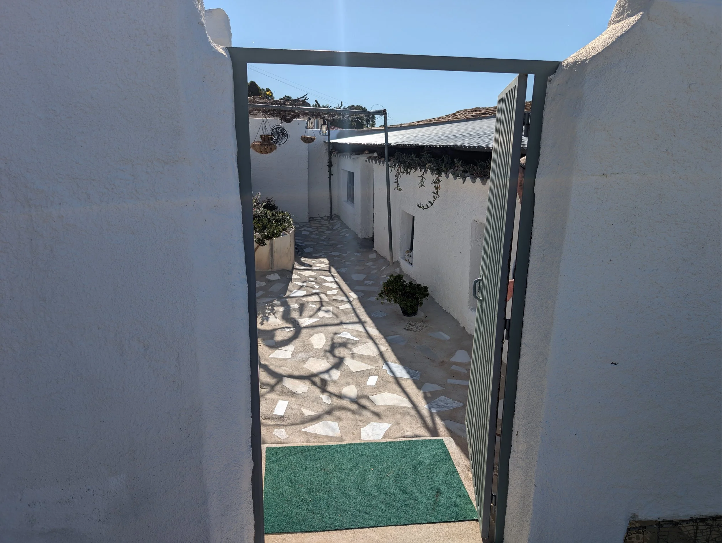 View through an open gate showing a small courtyard with plants, stone flooring, and white walls, with clear blue sky overhead.