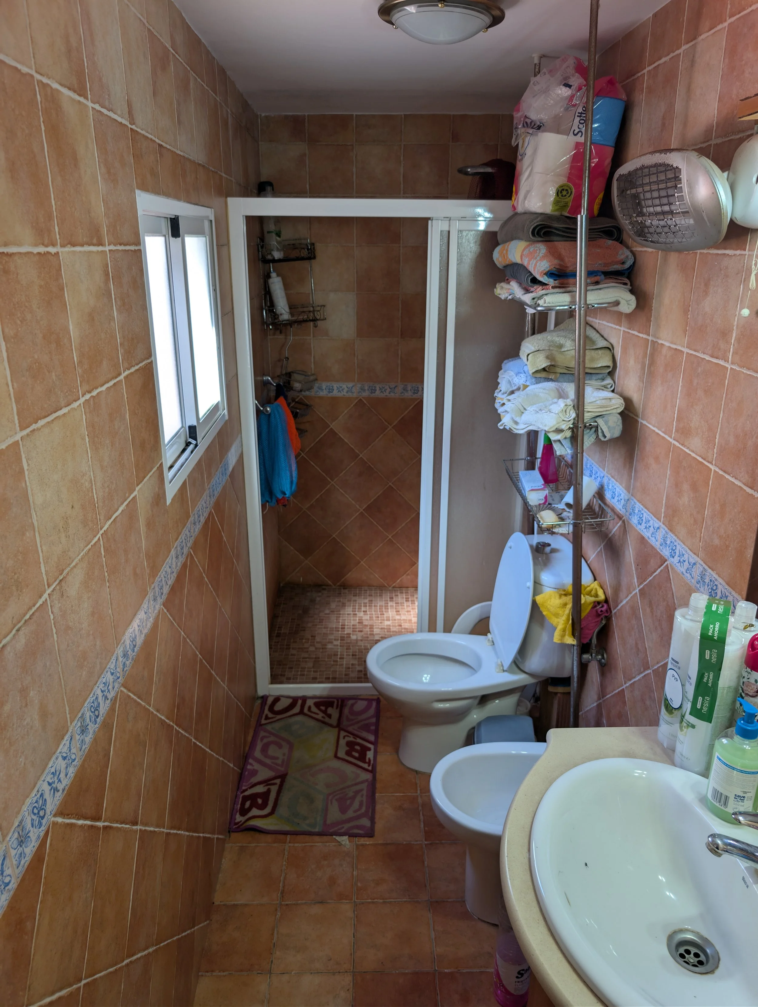 A small bathroom with brown tiled walls and floor, a window, a toilet, a bidet, a sink with toiletries, a rack with towels, and a walk-in shower with glass door and orange and brown tiles.