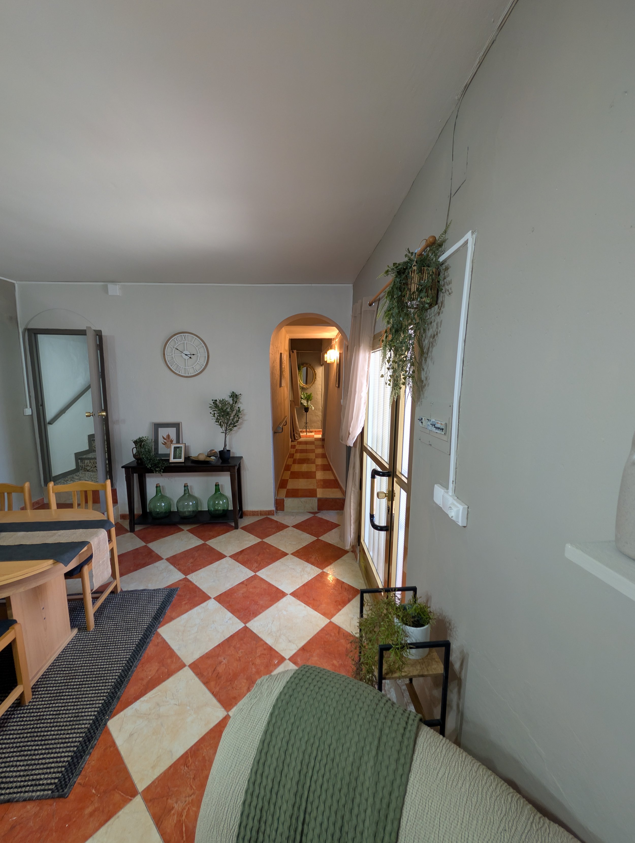 Interior view of a living room with tiled floor, a dining table with chairs, a console table with decorative items, and a hallway visible through an arched doorway. There are plants and wall decorations, including a clock, and a door leading outside.