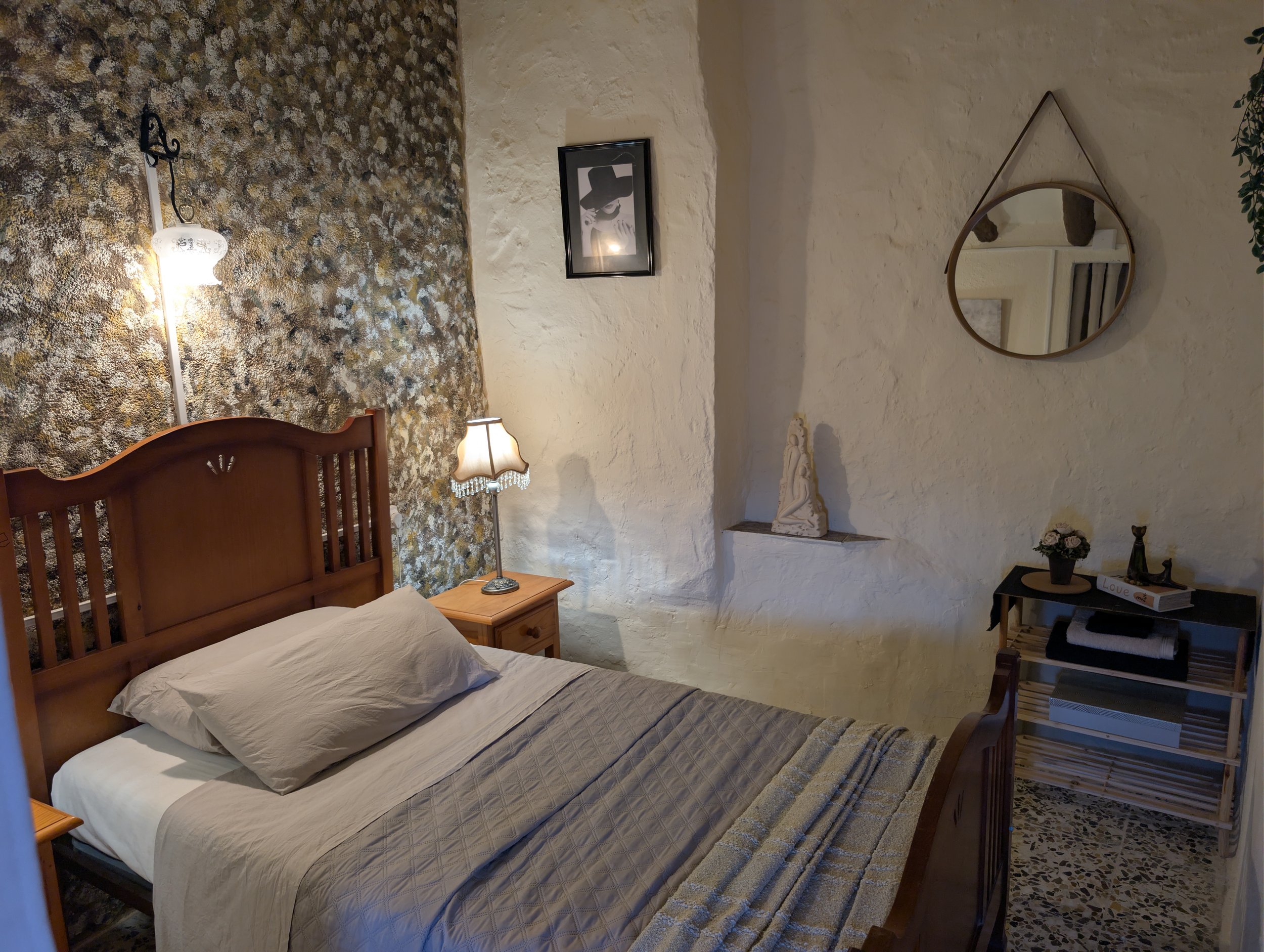 A cozy bedroom with a wooden bed, beige bedding, a small bedside table with a lamp, a wall-mounted framed picture, a mirror, a shelf with decorative items, and a textured white wall.
