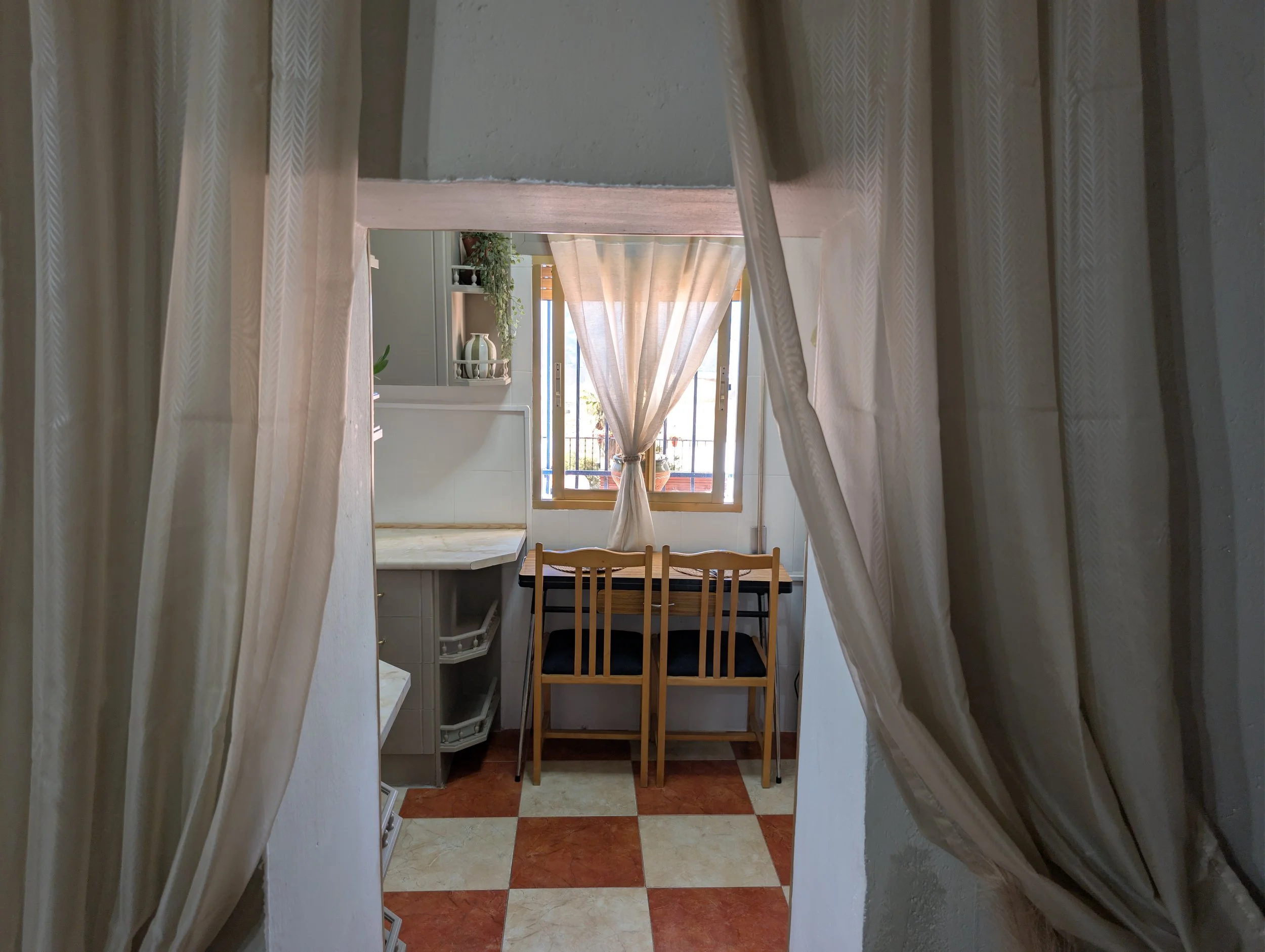 View of a small dining area with a window, beige curtains, and a wooden table with two chairs, seen through a doorway with cream-colored drapes.