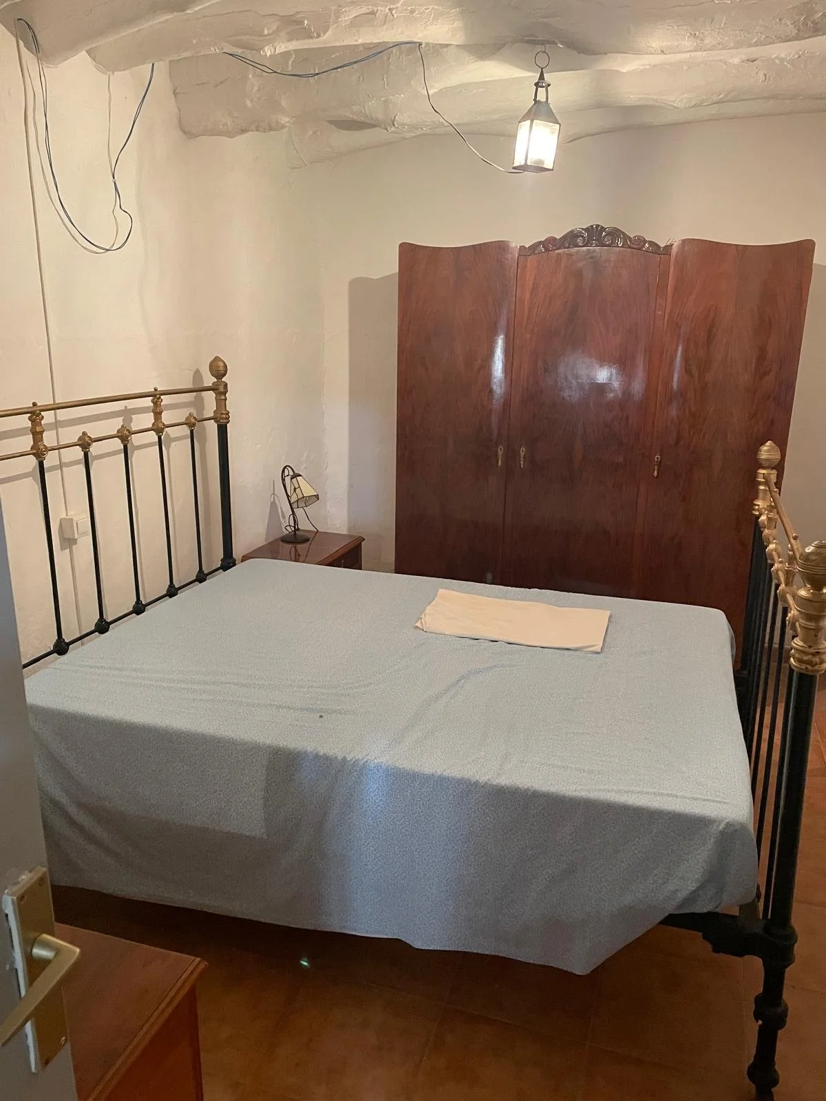 A bedroom with a metal bed frame, a mattress covered with a light-colored sheet, a blue towel on it, a small wooden nightstand with a lamp, a large wooden wardrobe, and a hanging lantern light from the ceiling.