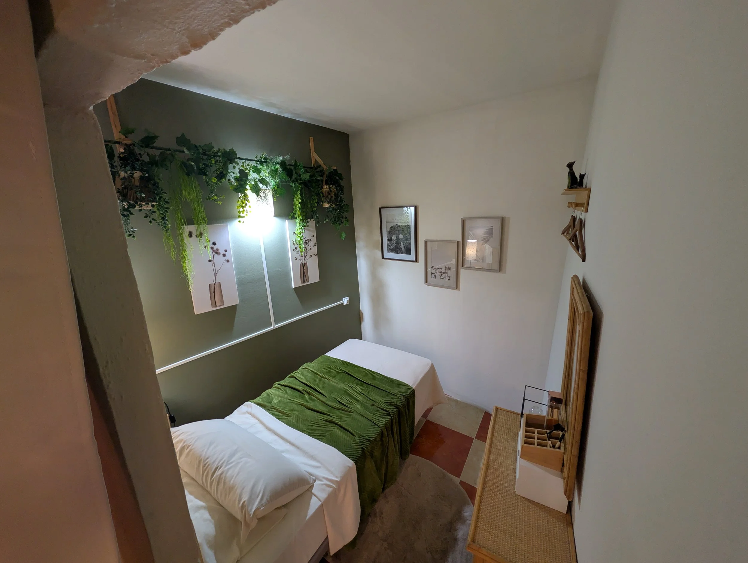 A small bedroom with a double bed draped in white linens, a green throw blanket, and a matching pillow. Above the bed, there's a green accent wall with decorative plant arrangements and artwork featuring vases with plants. On the right wall, three fr