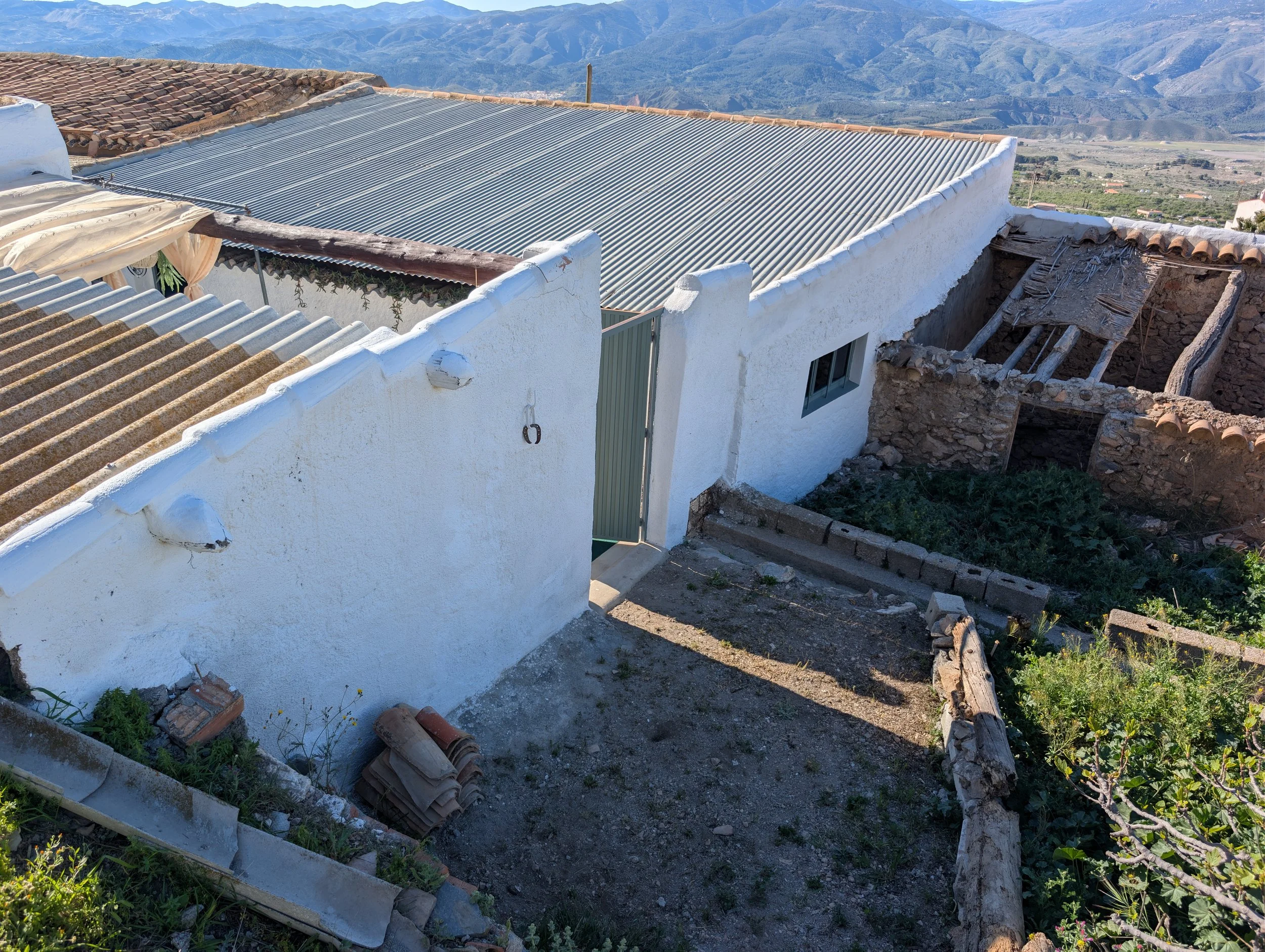Aerial view of a white building with a metal roof, damaged and collapsed section, and a closed green gate, set against mountain landscape.