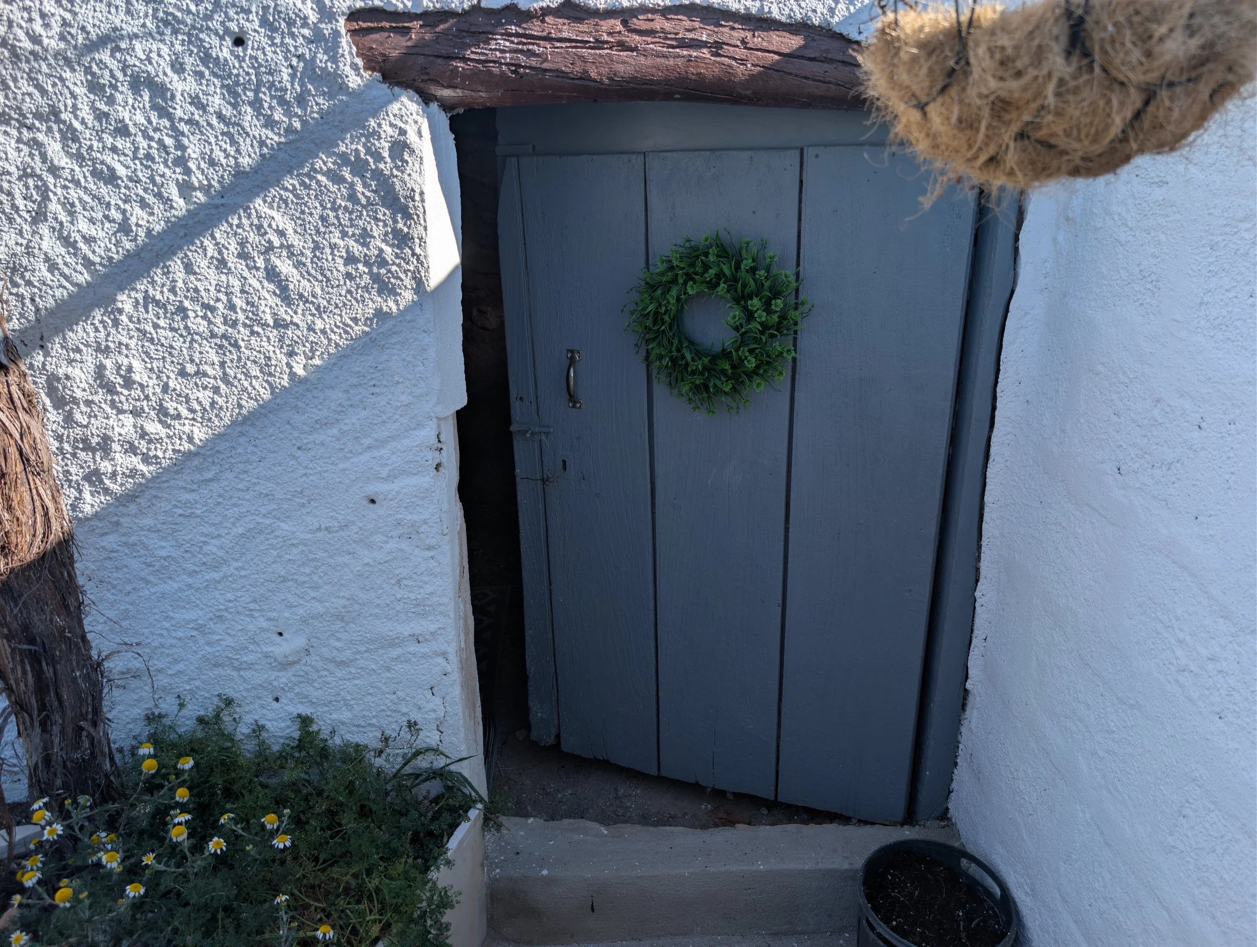 Gray wooden door with a green wreath, set between white textured walls, with a small plant on the ground and a black container nearby.