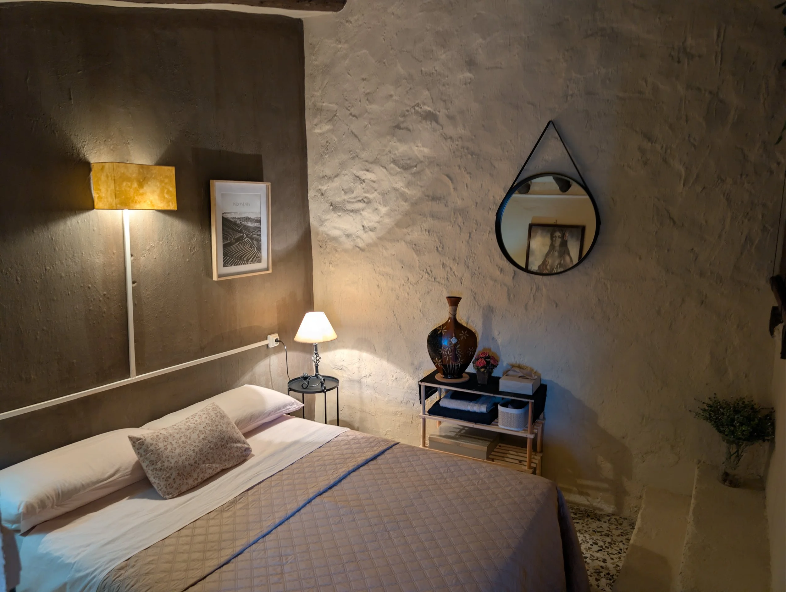 A cozy bedroom with a beige bedspread, pillows, a small black side table with a lamp, a wall mirror, a decorative vase on a small shelf, framed pictures on the walls, and potted plants.