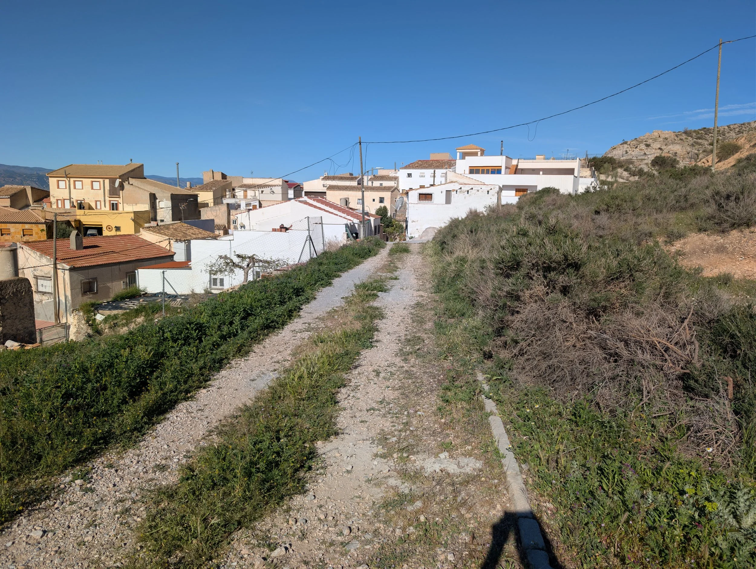 A gravel pathway leading towards a small village with white and beige houses on a hillside under a clear blue sky.