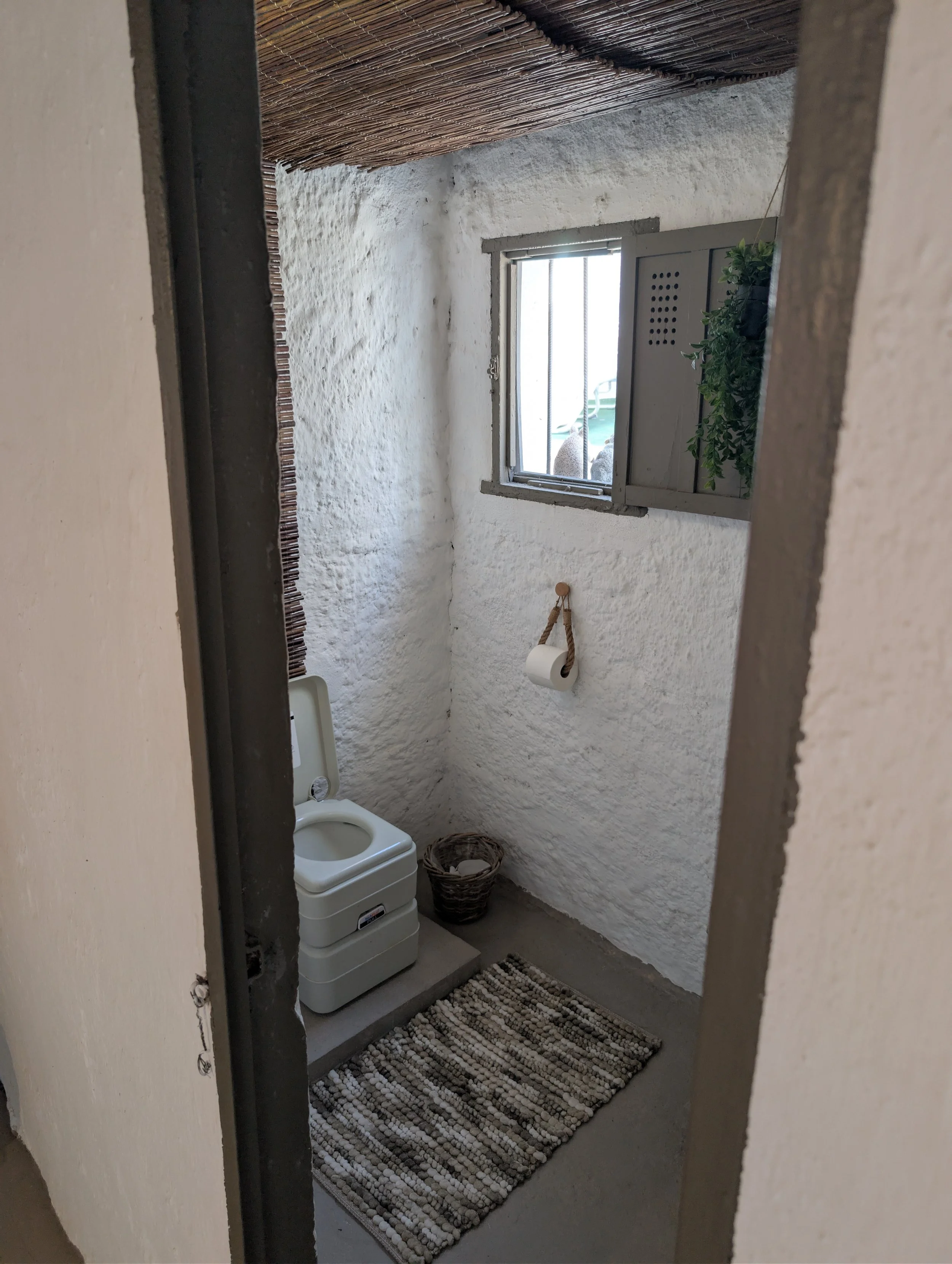 Small rustic bathroom with white textured walls, a small window with bars, a portable toilet in the corner, a woven basket on the floor, a paper towel holder, and a rug on the concrete floor.
