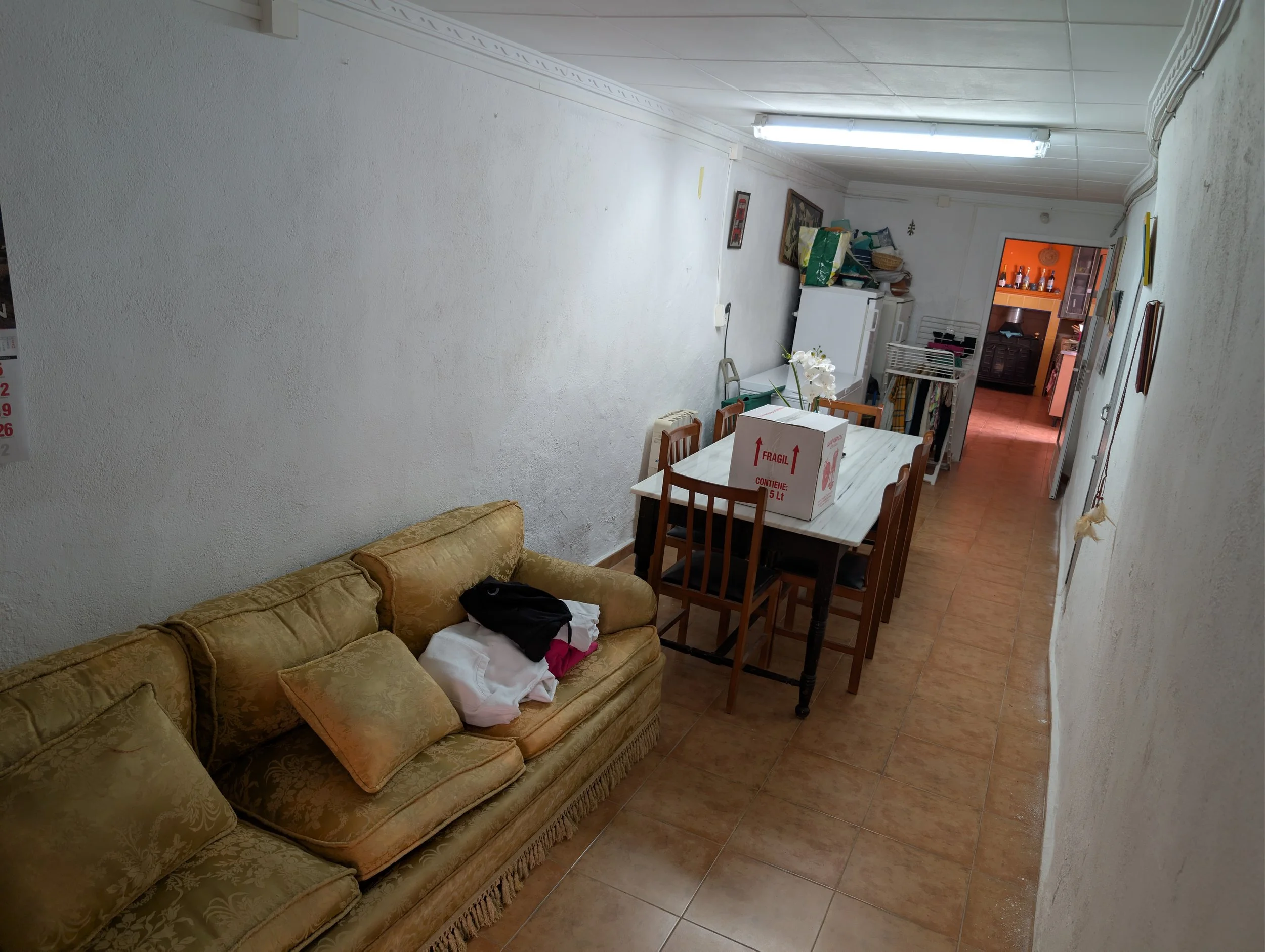 A room with a vintage yellow sofa, a wooden dining table with six chairs, and a large cardboard box on the table. The room has tiled floor, white walls, and a fluorescent ceiling light. In the background, there is a white refrigerator, kitchen space,