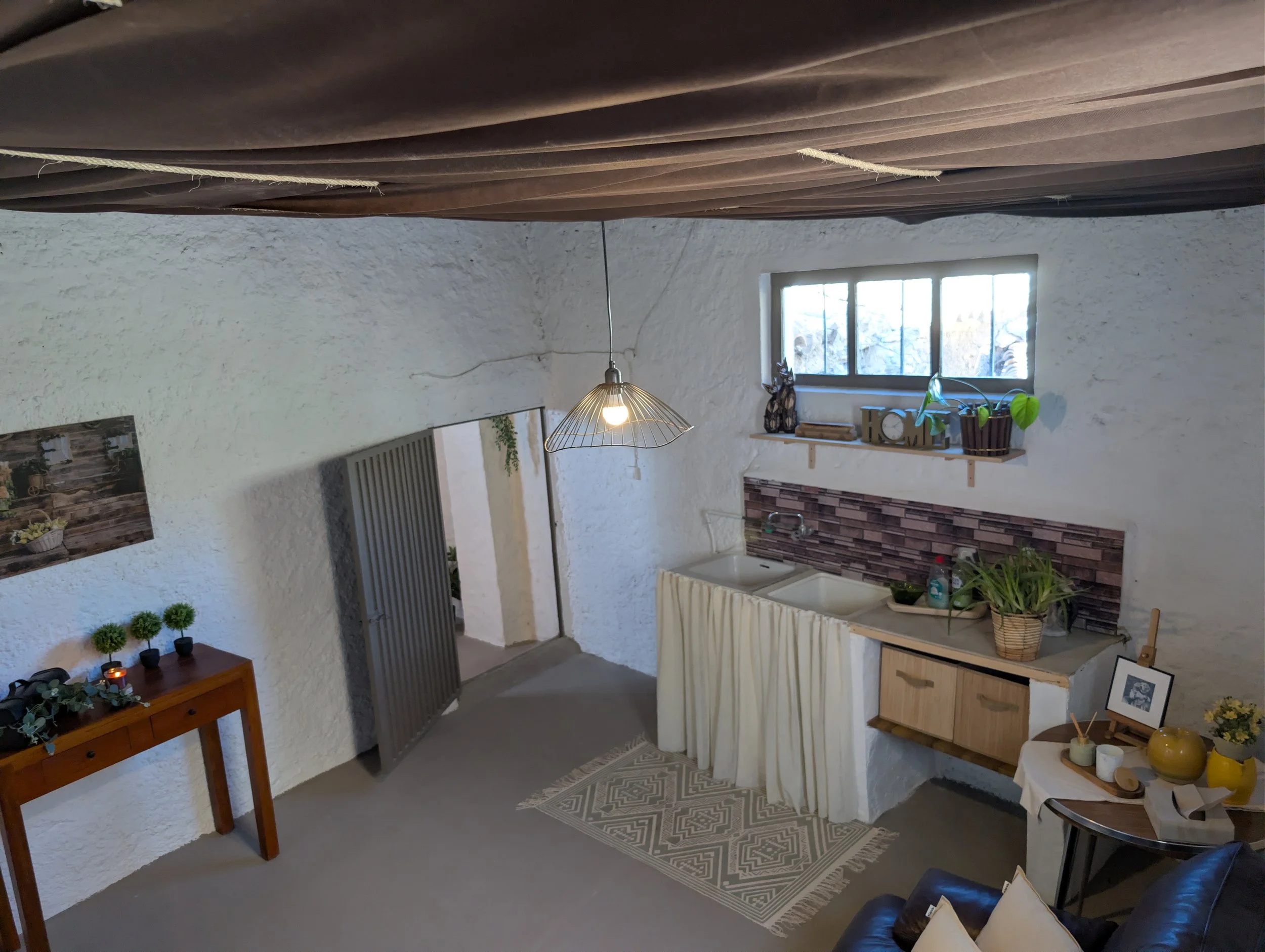 Interior of a rustic kitchen with white textured walls, a small window with plants on the windowsill, a wooden cabinet with plants and decorations, a striped rug, and a round table with various decor items.