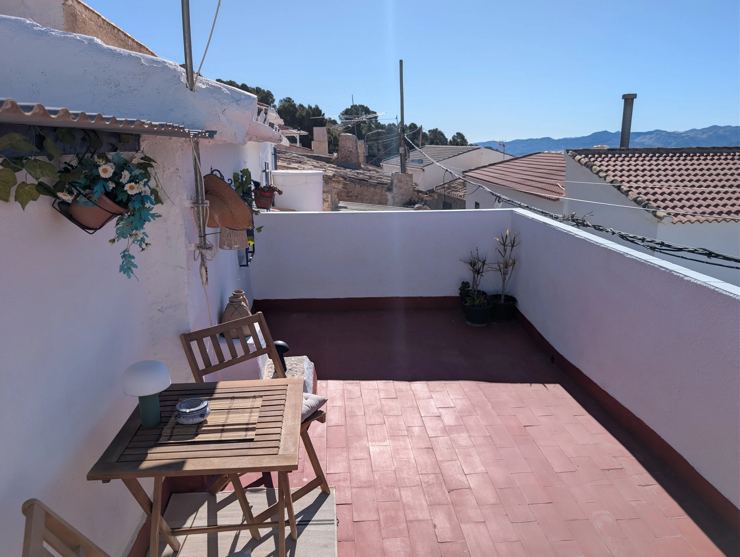An outdoor terrace with a pink tiled floor, white walls, a wooden table and chairs, potted plants, hanging flower baskets, and a traditional straw hat hanging on the wall under a sunny blue sky with mountain views in the distance.