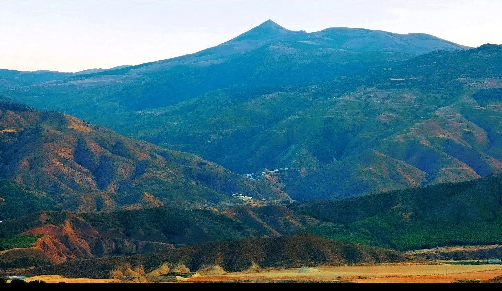 Mountain landscape with green and brown hills and a tall mountain peak in the background.
