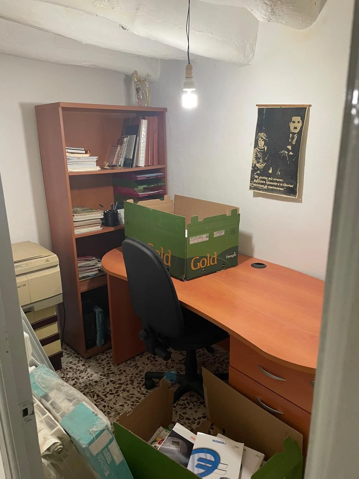 A small office room with a wooden desk, black office chair, and a wooden bookshelf filled with books and files. There is a cardboard box labeled 'Gold' on the desk and another box on the floor with various items. The room has a single light bulb hang