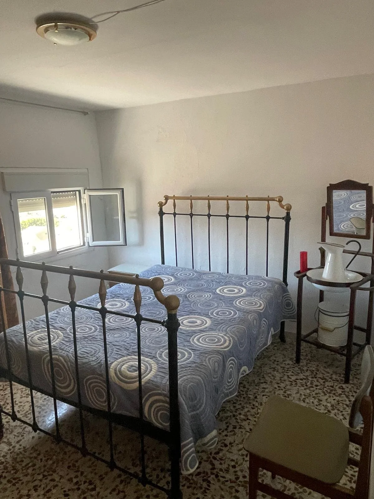 A bedroom with a metal bed frame, a window partially open, a small mirror, a chair, and a small table with a white pitcher, red candle, and other items. The floor has a speckled tile pattern, and the walls are white.