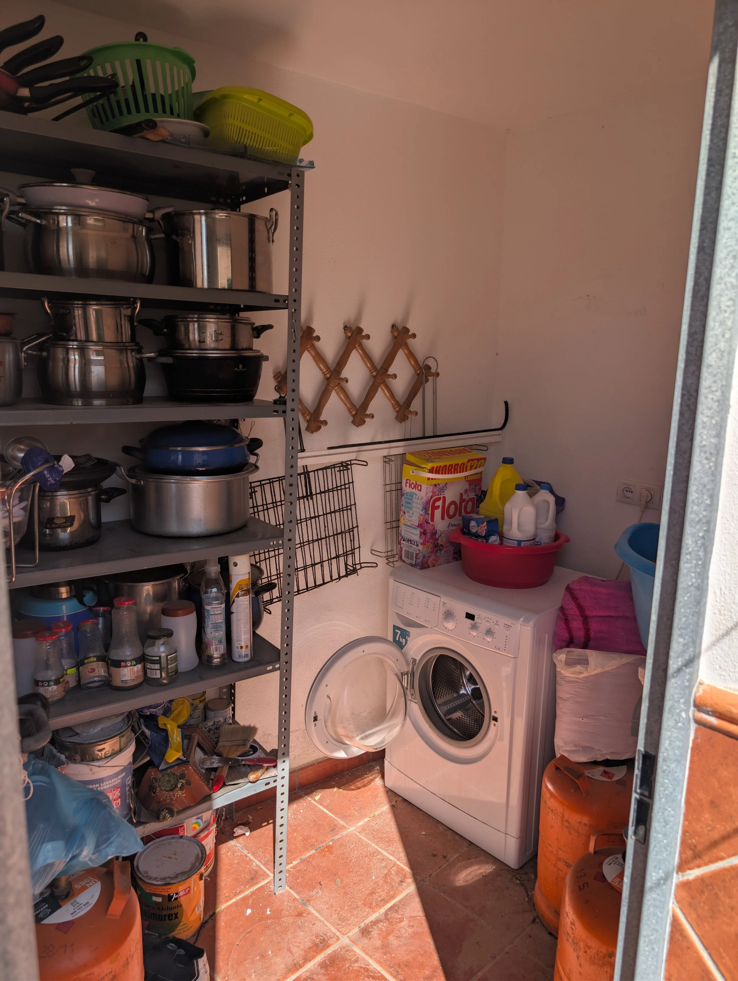 A laundry room with shelves holding pots, jars, and containers. There is a washing machine with the door open, laundry supplies on top, and several orange propane tanks on the floor. Pink towels and a large plastic basin are also present.