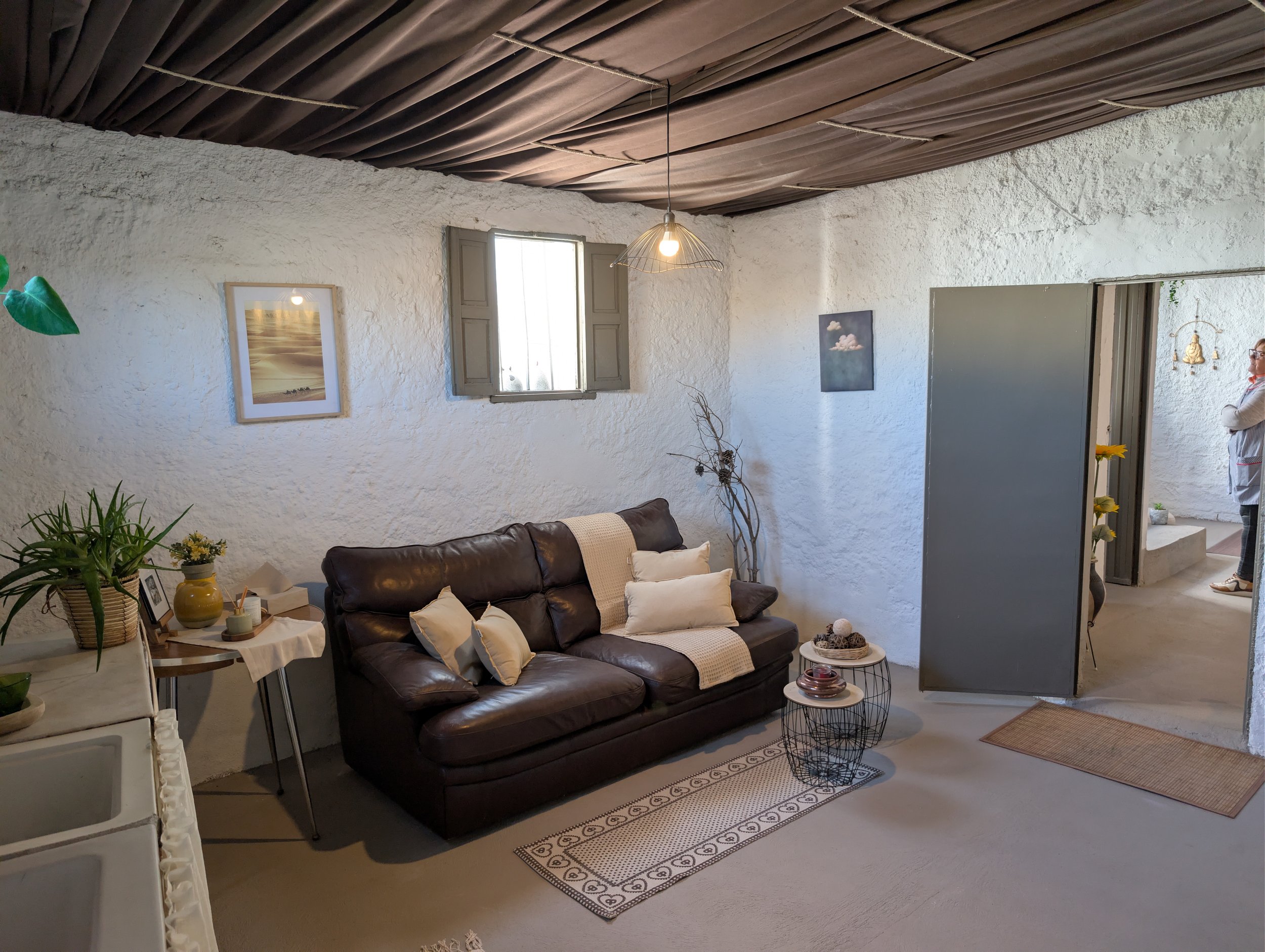 Cozy living room with white textured walls, a dark brown leather sofa with beige and white cushions, a small side table with decorative items, a potted plant, framed artwork on the walls, and a doorway leading to another room. The ceiling has dark dr