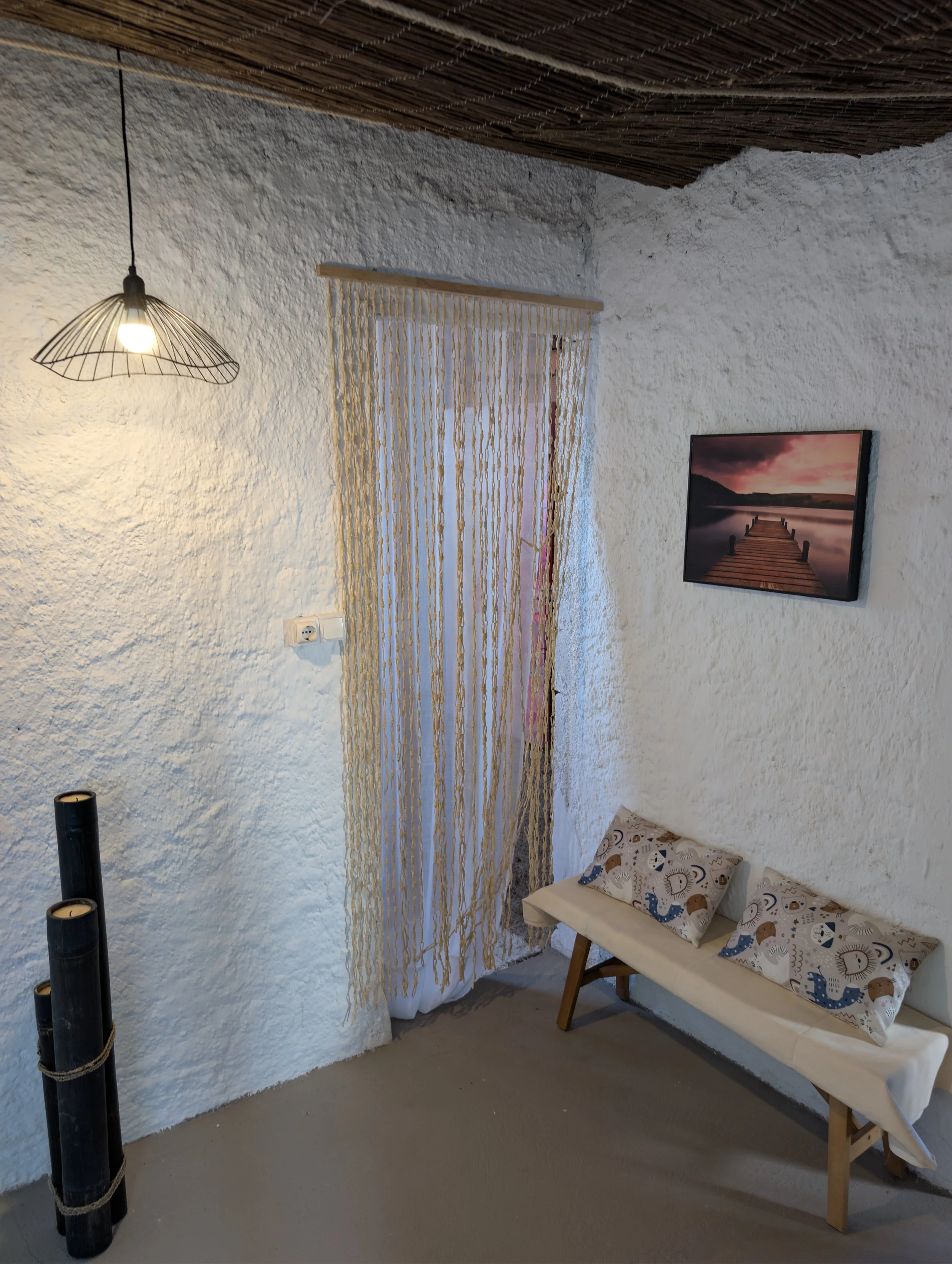 Interior corner of a room with white textured walls, a hanging wire-frame light, a beaded curtain, a landscape photograph of a pier over water at sunset on the wall, a small wooden and cushioned bench with animal-themed pillows, and three tall black 