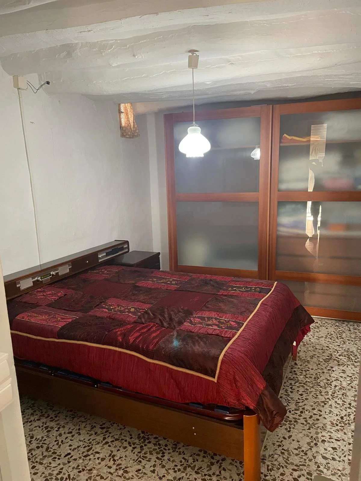 A bedroom with a double bed covered by a red and brown patterned quilt. There is a nightstand on the left side of the bed, a hanging ceiling light, and a sliding door with frosted glass panels at the back.