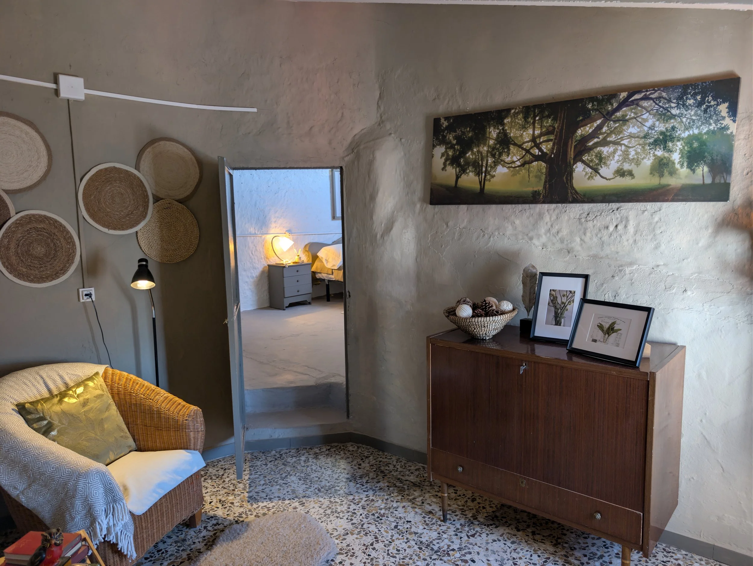 A cozy interior room with a wicker chair with a yellow pillow, a wall decorated with round woven baskets, a black floor lamp, a wooden cabinet with framed pictures and a basket of decorative balls, and a large landscape painting of trees on the wall.