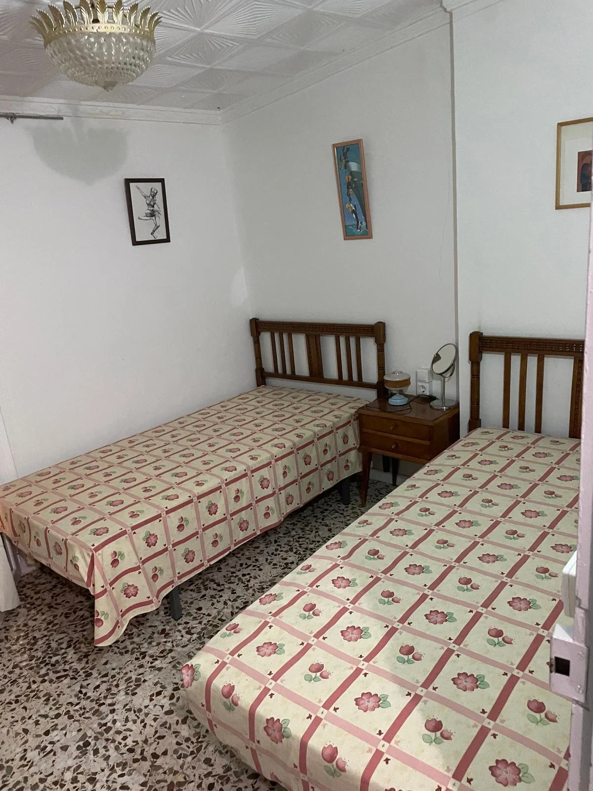 A bedroom with two single beds with matching floral bedspreads, wooden headboards, a wooden nightstand with a lamp and mirror between the beds, white walls decorated with framed art, a white ceiling with a decorative light fixture, and a terrazzo flo