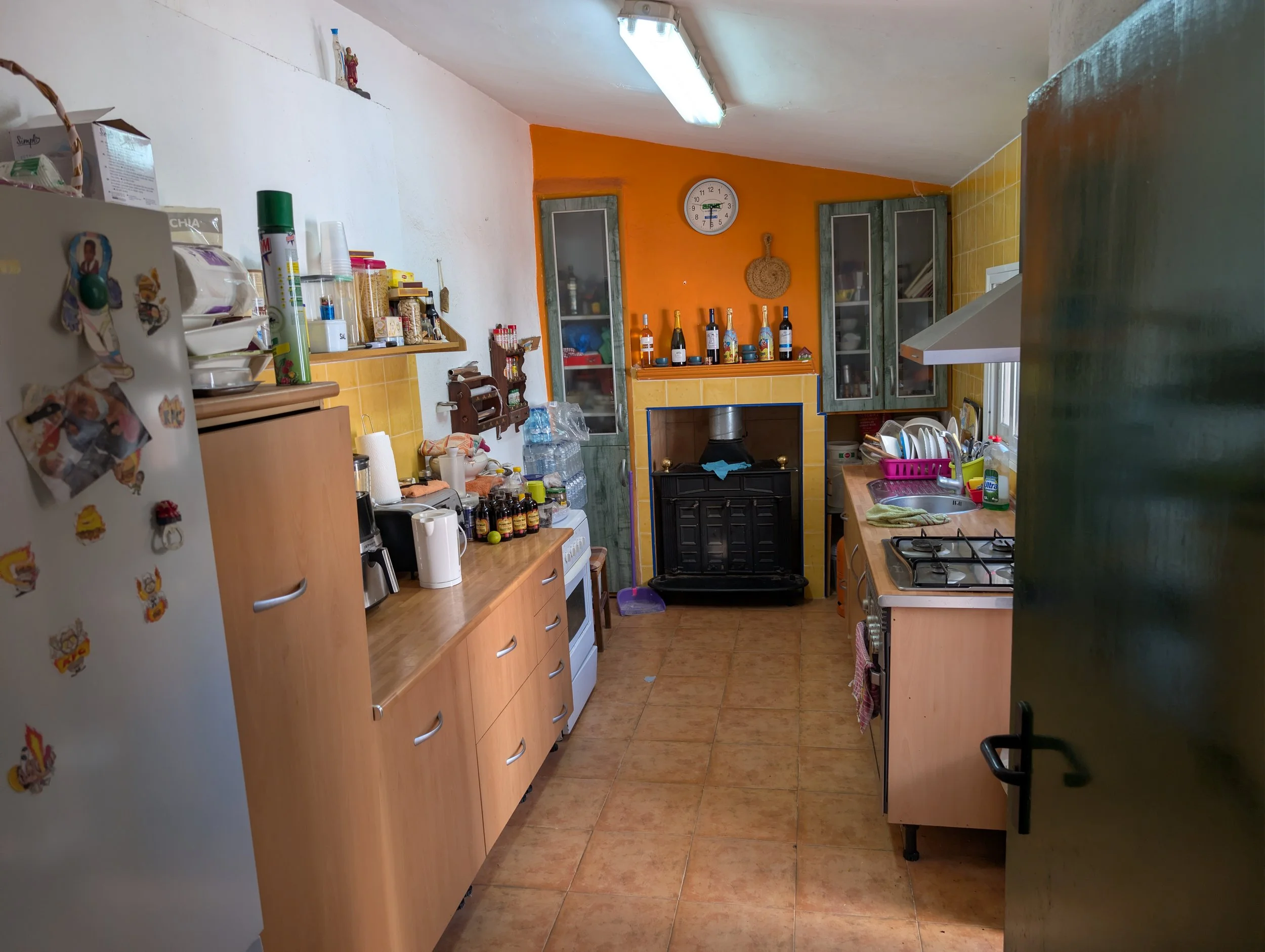 A small kitchen with beige cabinets, a white refrigerator adorned with stickers, and various kitchen appliances. The walls are painted white and orange. Items like water bottles, bottles of alcohol, dishes, pots, and kitchen utensils are on the count
