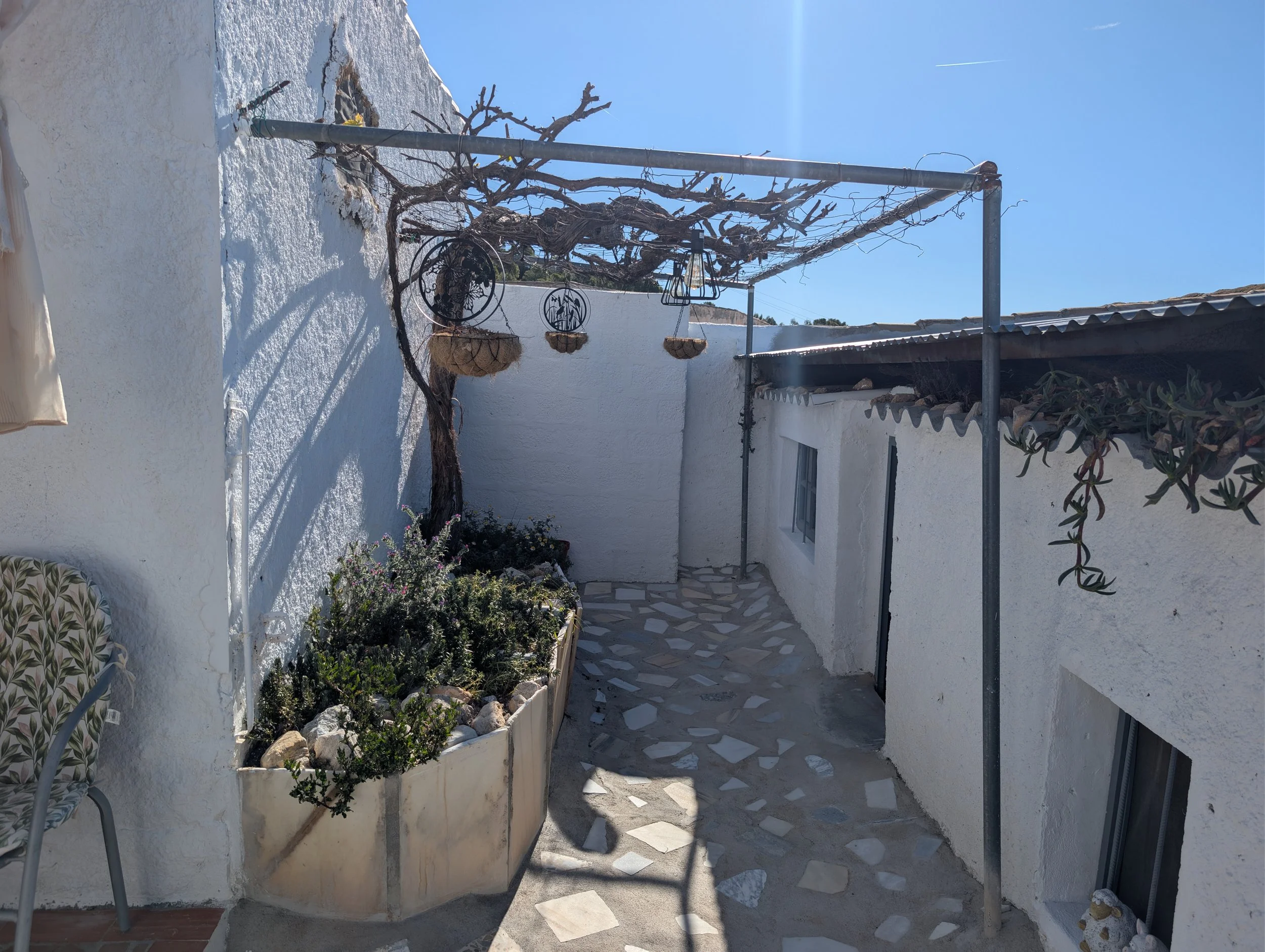 Small outdoor patio with white textured walls, a vine growing on a metal frame, and hanging baskets. Potted plants and a chair are also present.