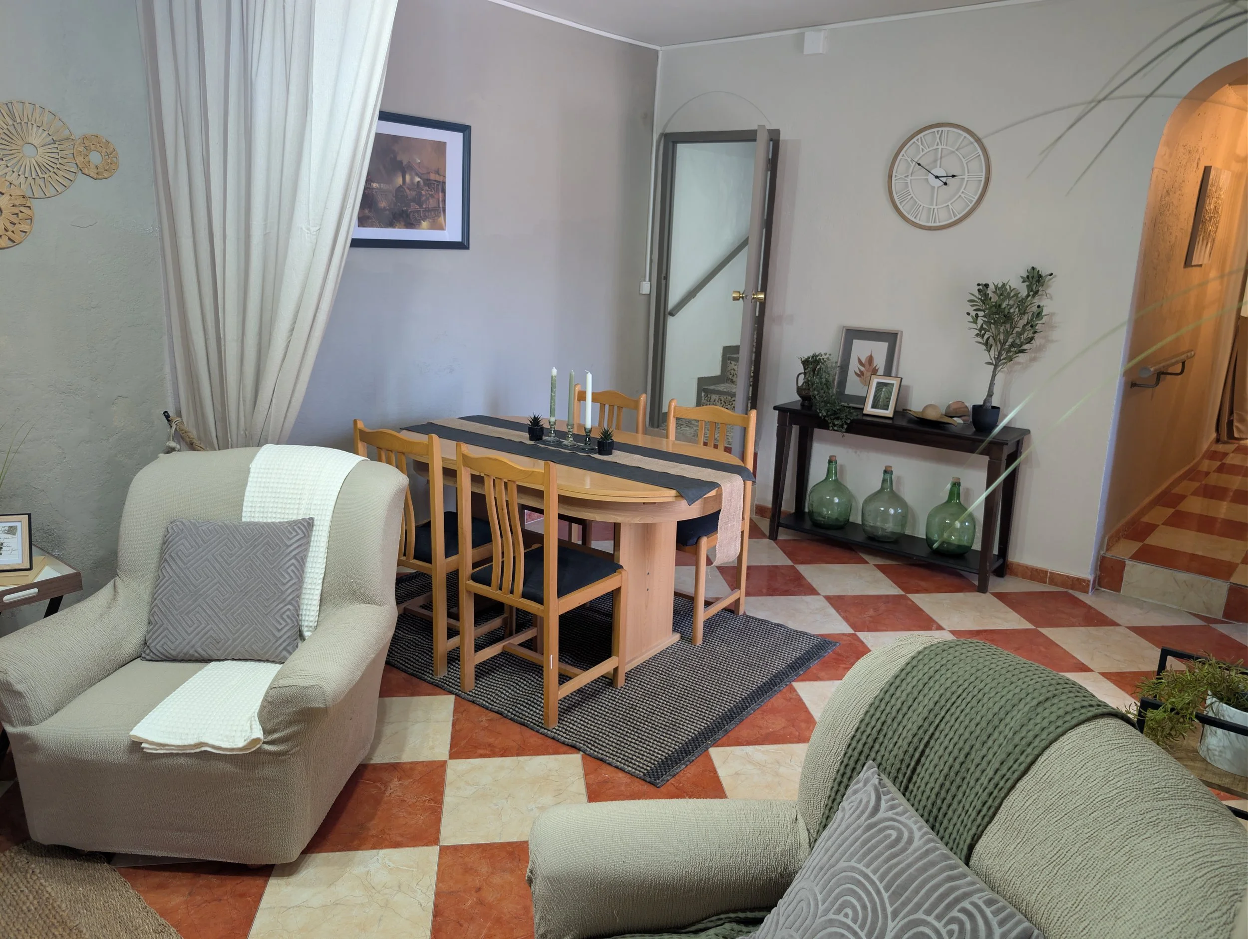 Living room with beige and green armchairs, a wooden dining table with six chairs, a black console table with decorative vases and framed pictures, a wall clock, a mirror, and a picture on the wall. The floor has a checkered pattern of red and cream 