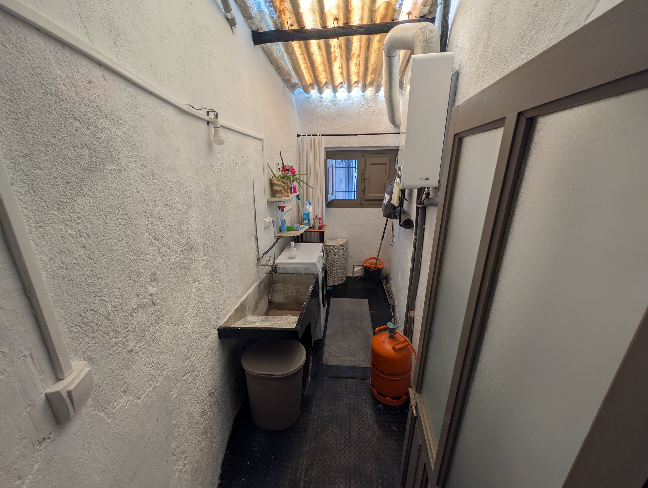 Small laundry room with white textured walls, a black rubber floor, a utility sink, a gray trash can, supplies on a small shelf above the sink, a window with bars, and a gas cylinder.