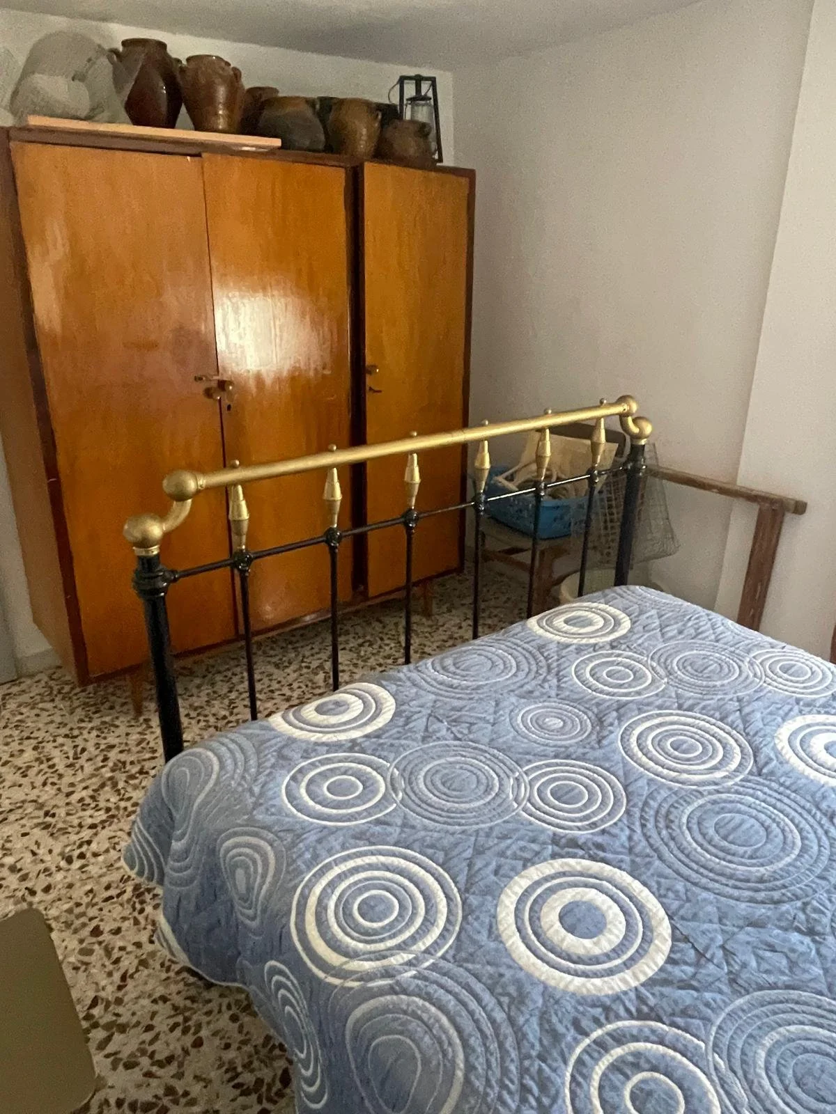 A bedroom with a brass and black metal bed frame, a mattress with a blue and white circular patterned quilt, a wooden wardrobe with brown pottery on top, and a small table with a blue laundry basket and a fishing net on it.