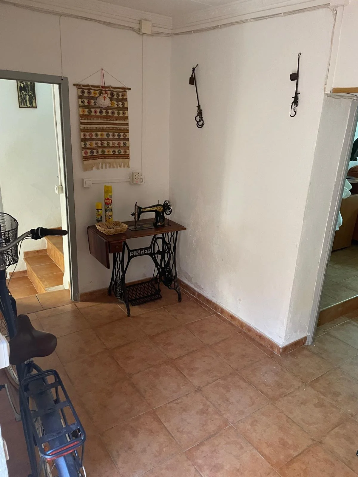 A corner of a room with a vintage black sewing machine on a small wooden table, two cans of spray, a small basket, and a decorative fabric wall hanging. Two hooks are hanging from the wall, and a staircase with brown steps is visible through a doorwa